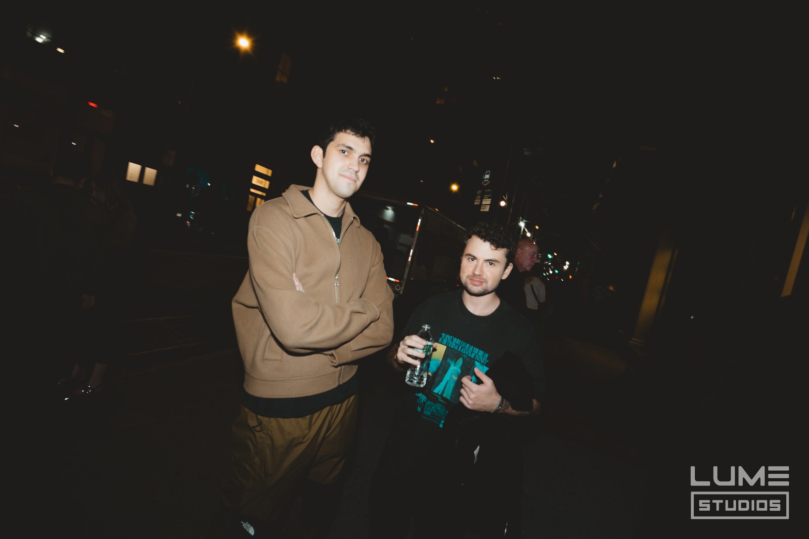 Two men pose on a night street, one in a tan jacket, the other holding a green can