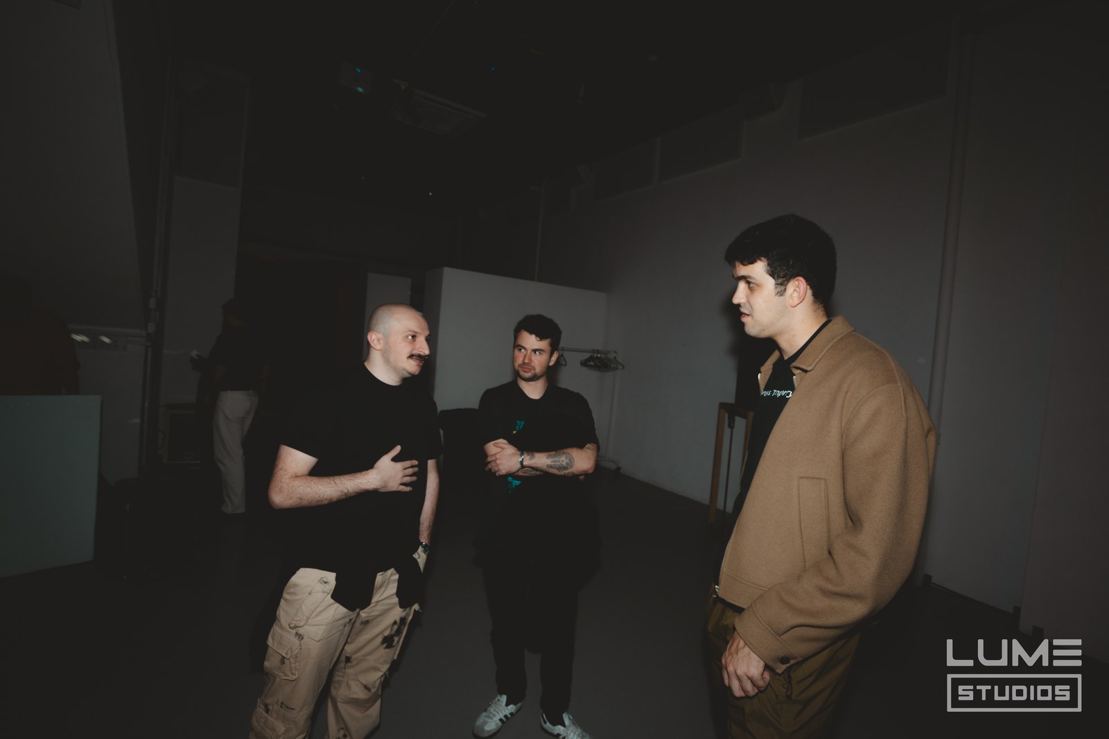 Three men in conversation inside a dim studio, one in a tan jacket, one gesturing in a black tee