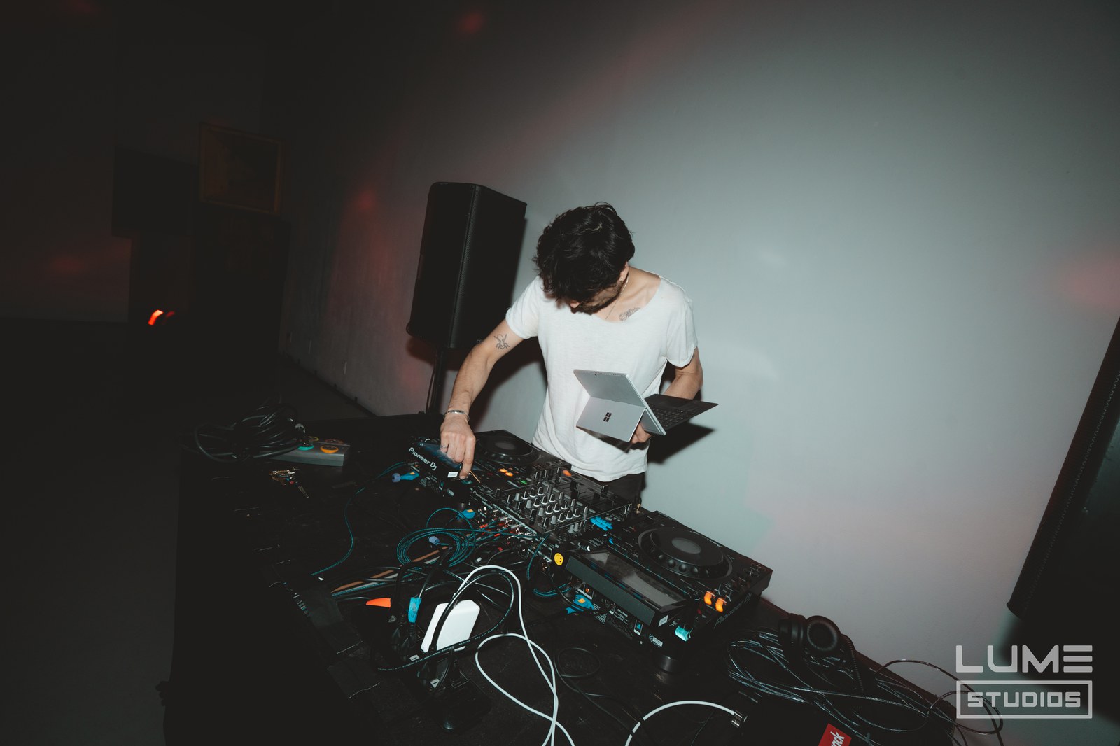 DJ in a white tee bends over a mixer and tangled cables beside a tall speaker under dim red light