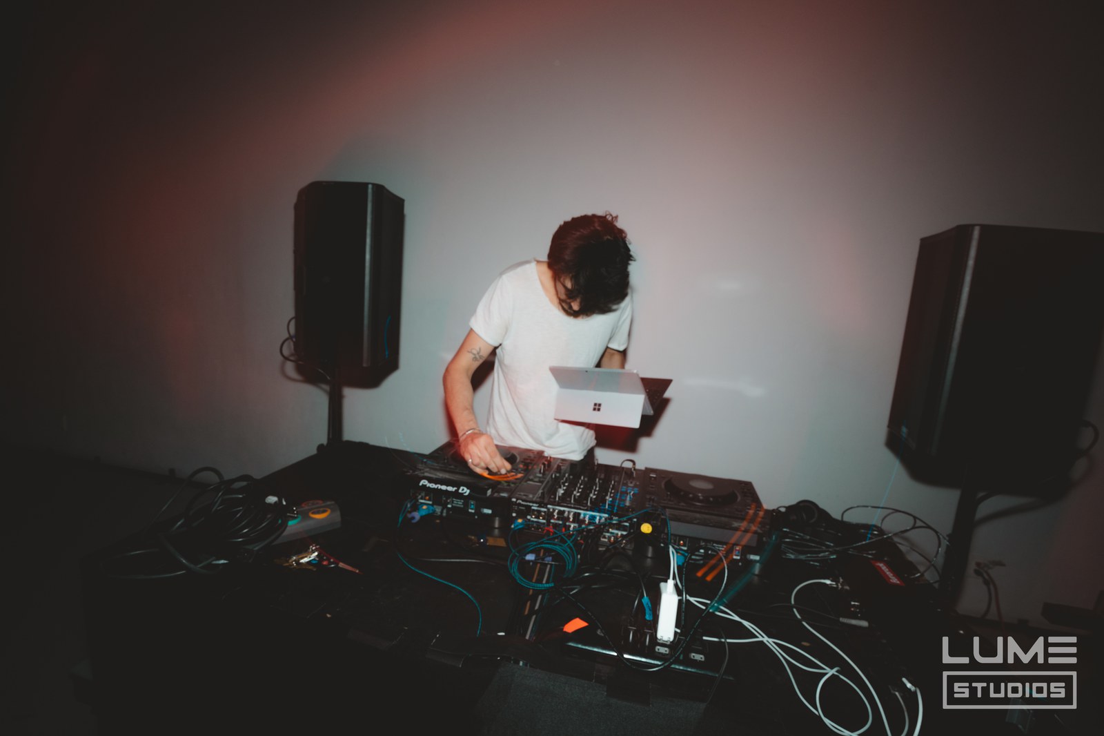 DJ in a white tee leans over a mixer and laptop between two tall speakers under pink light