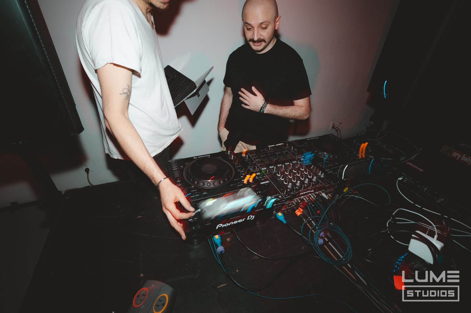Two DJs work a Pioneer mixer and CDJ under pink light, one pressing a pad with headphones nearby