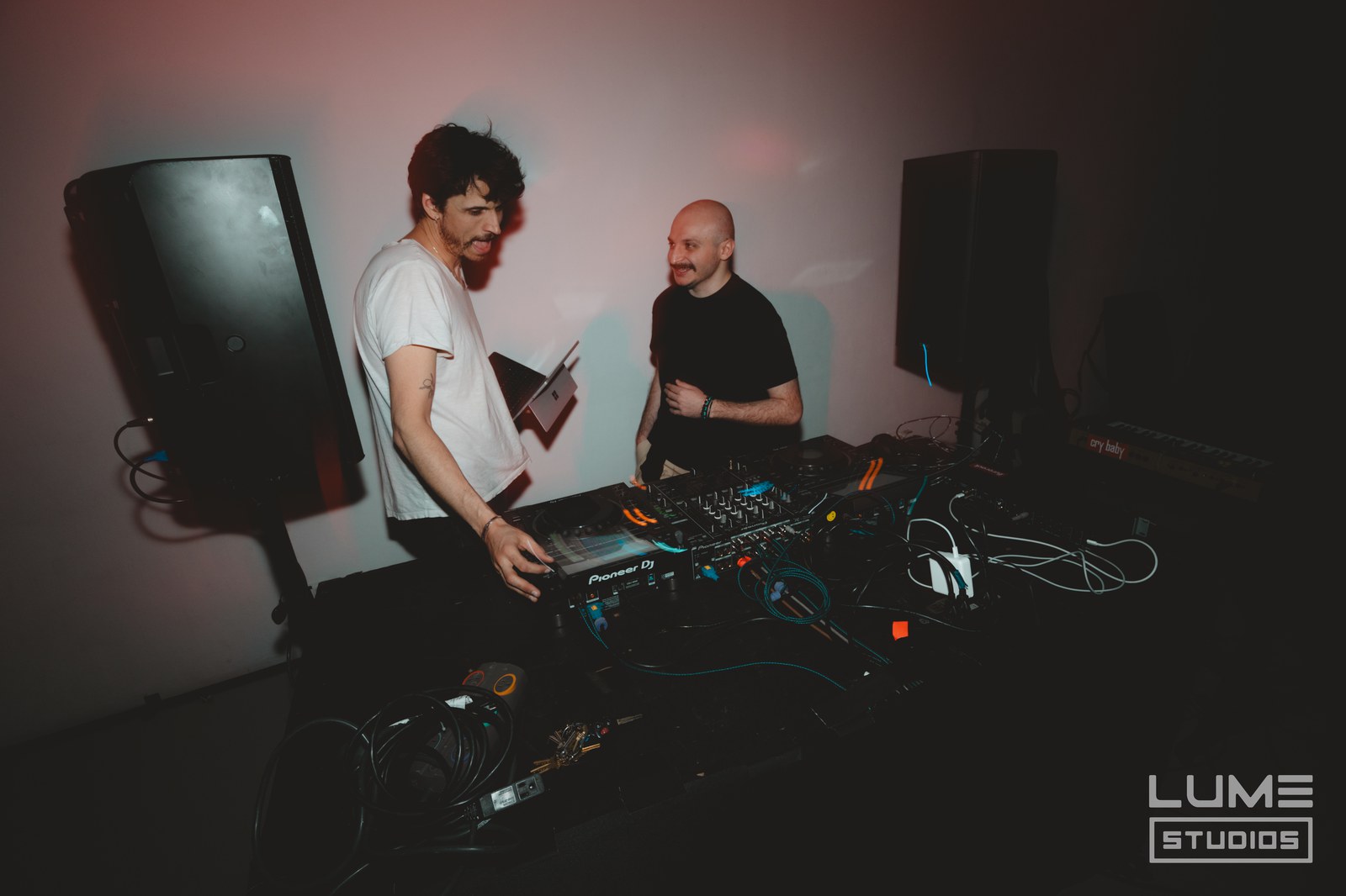 Two DJs share a laugh behind a Pioneer mixer flanked by speakers in a dim room