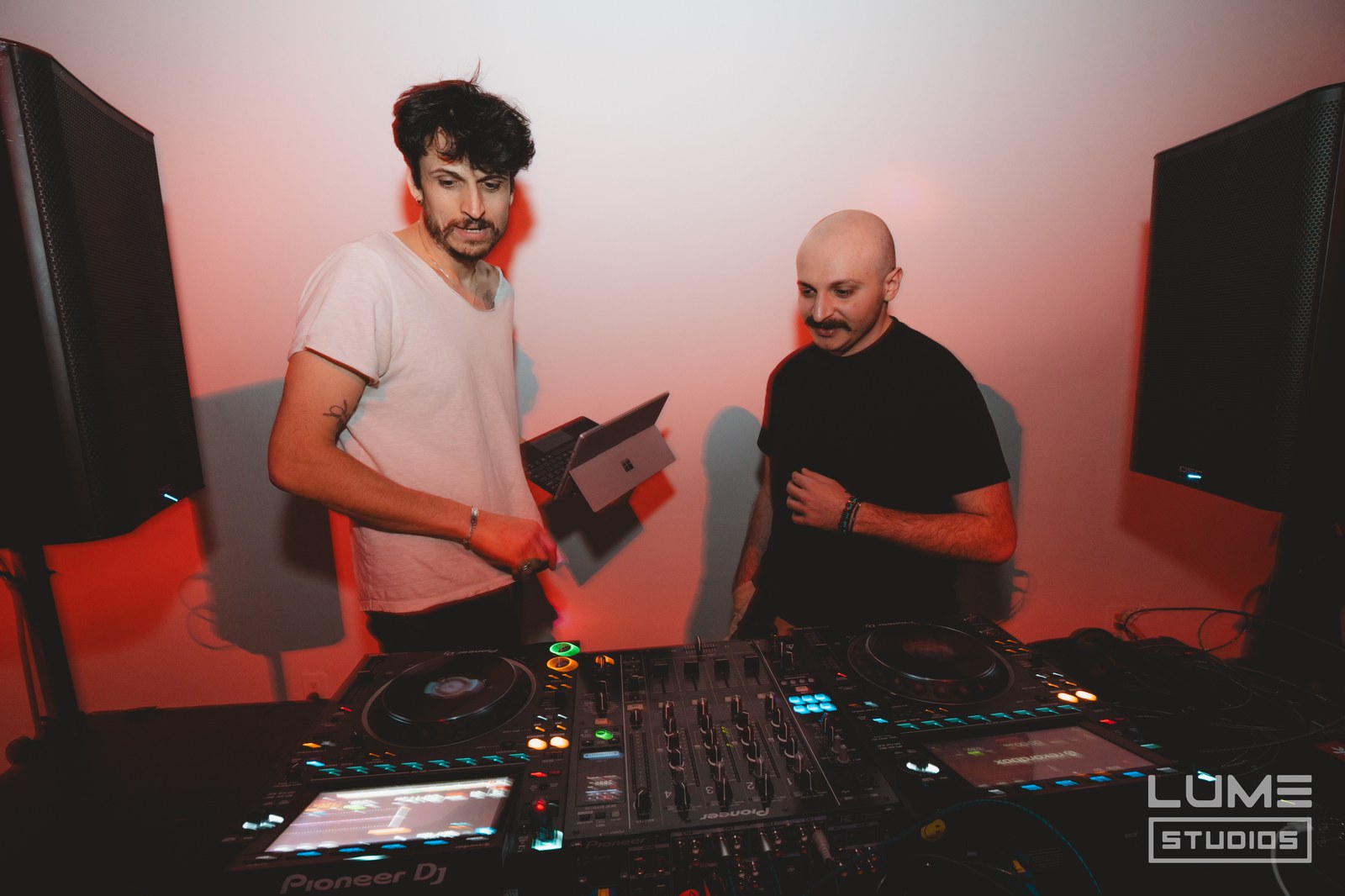 Two DJs behind Pioneer decks, one in a white tee and one in black, against a red lit wall