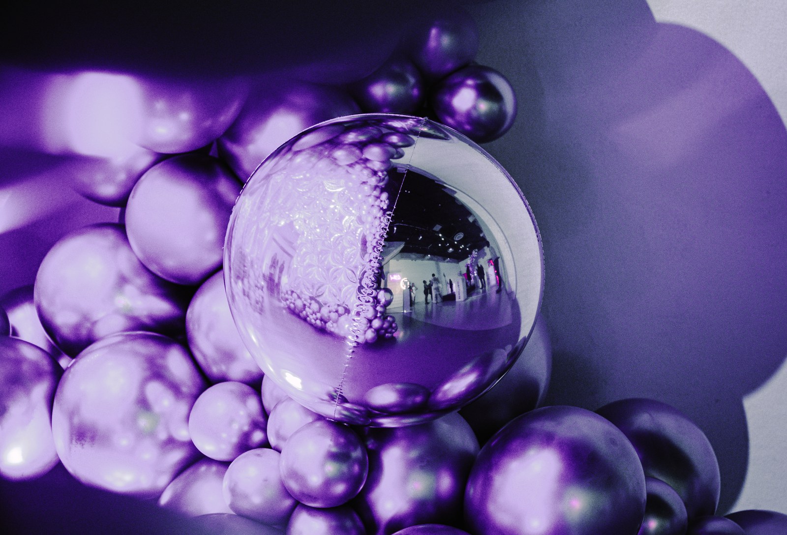 A cluster of metallic purple balloons with one clear mirrored sphere reflecting a lit room and guests