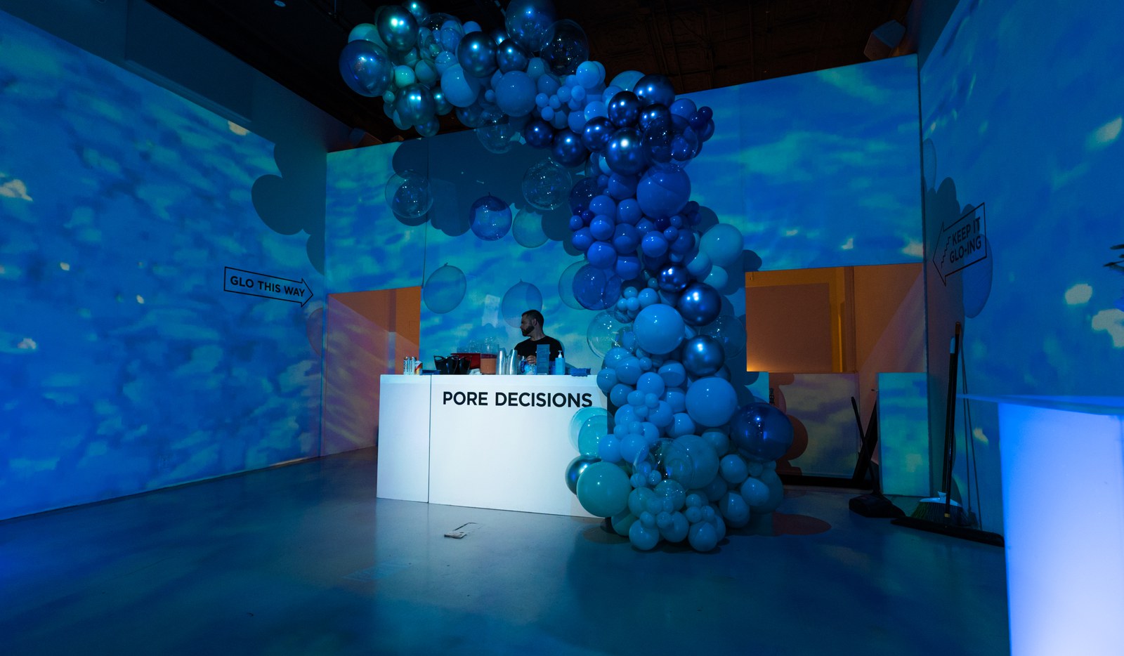 A DJ plays behind a white booth reading Pore Decisions, wrapped in a cascade of blue balloons under watery projections