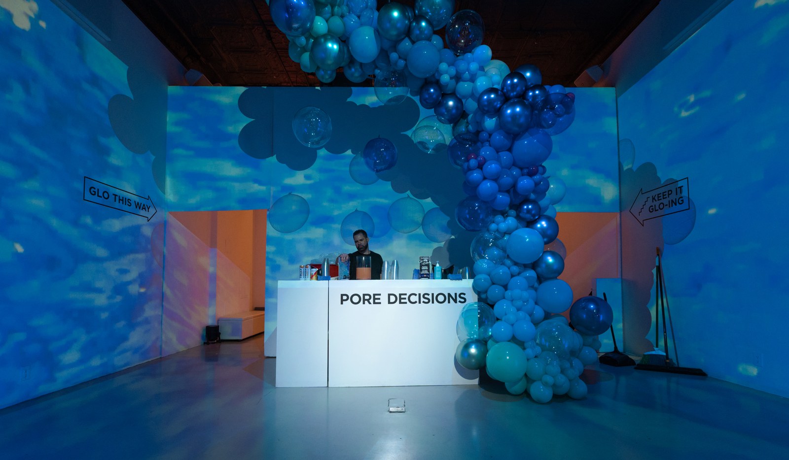 A bartender stands behind a white bar labelled Pore Decisions beside a cascade of blue balloons under water light projections
