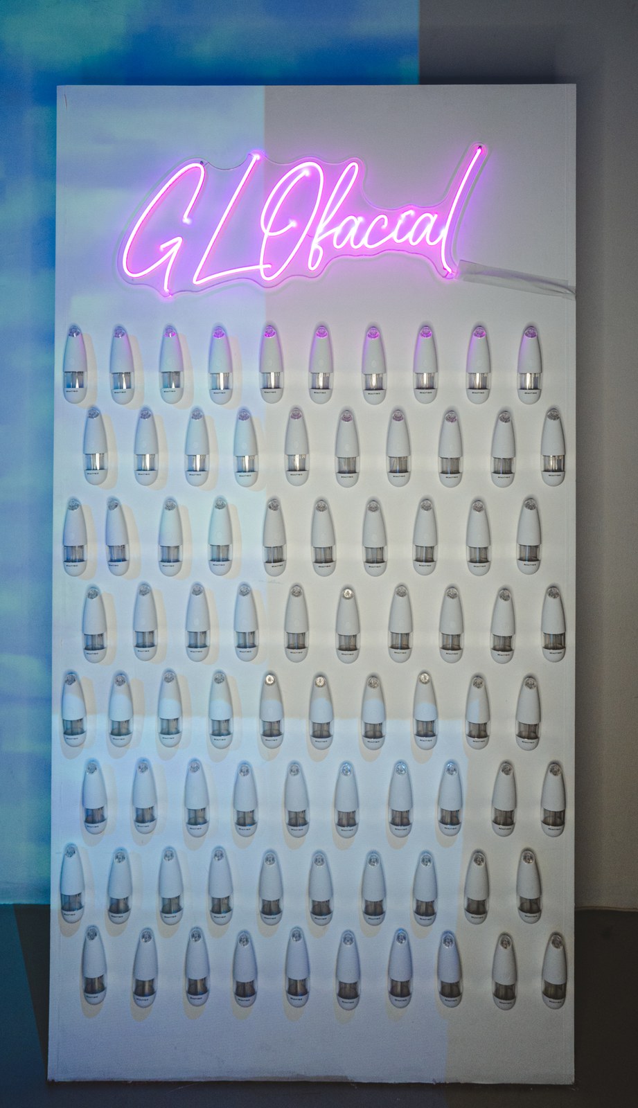 White wall of rows of GLOfacial handheld devices beneath a pink neon script sign