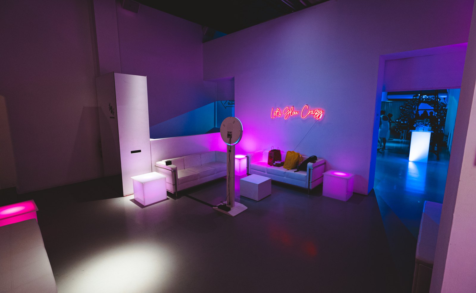 A pink lit lounge with a white sofa, glowing cube stools, a ring light and a neon sign