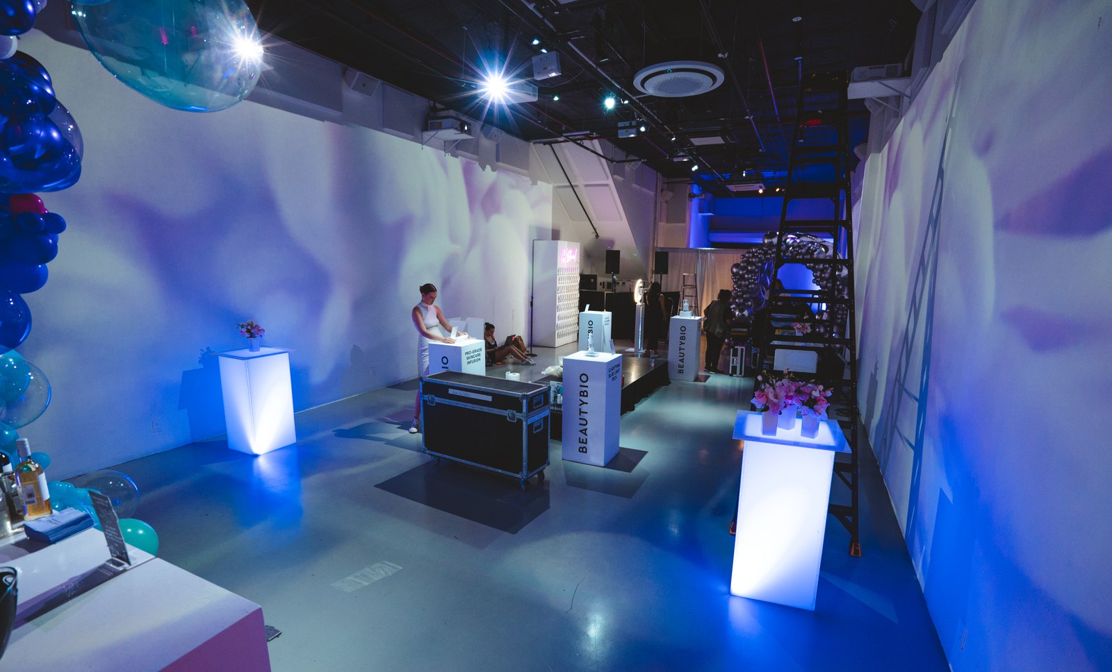 Blue lit room with glowing white plinths, a BeautyBio display and balloons beside a lone attendant