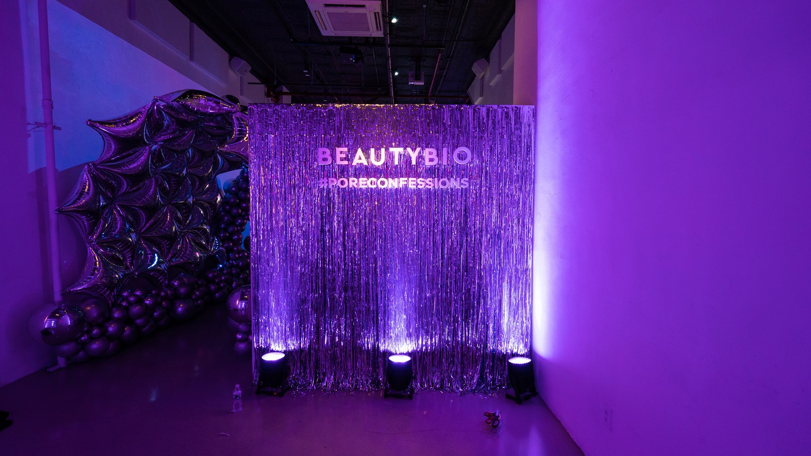 A silver tinsel curtain printed with the BeautyBio name stands beside metallic balloons in a purple lit hallway