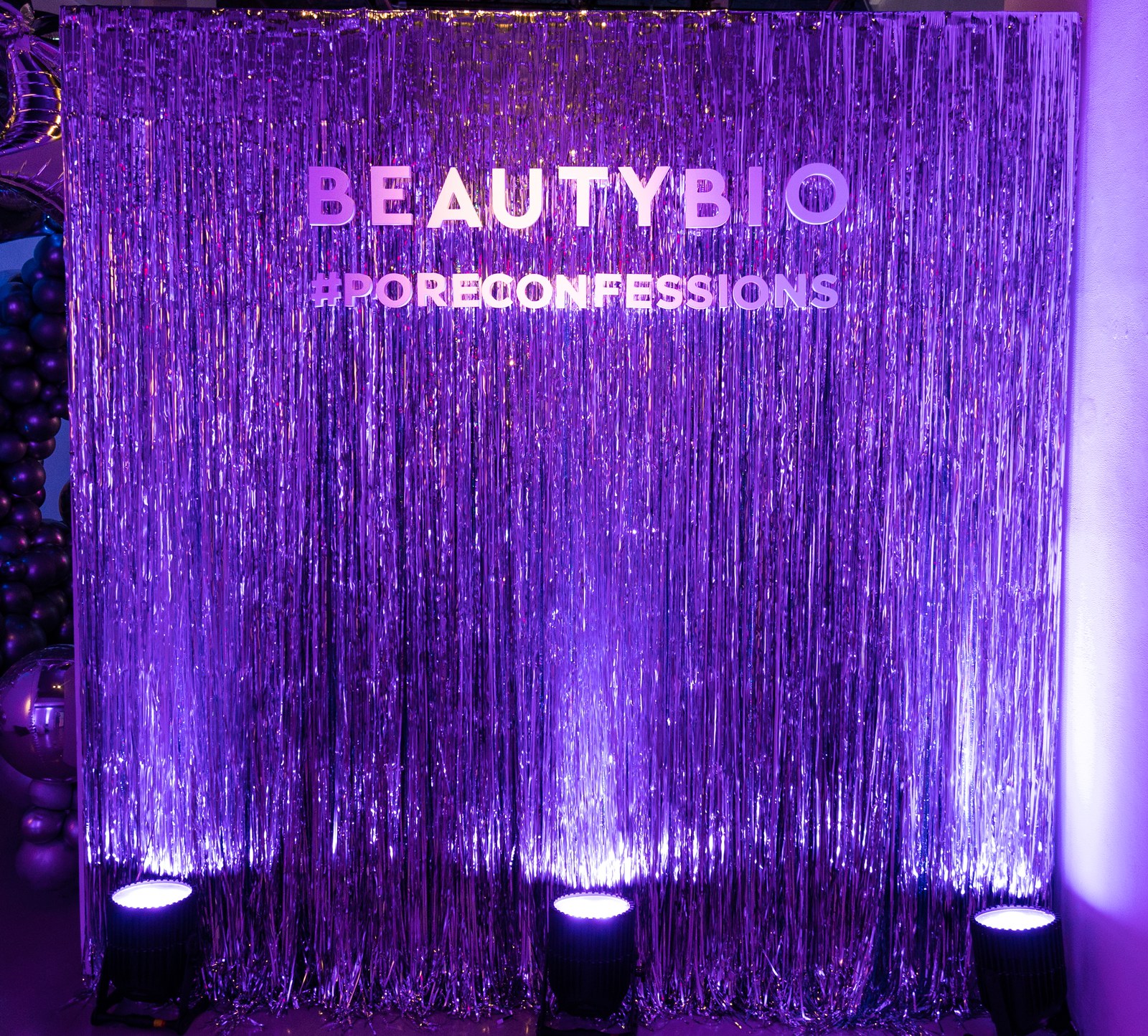 Purple lit silver fringe curtain backdrop with BeautyBio and #PoreConfessions lettering, three uplights along the floor