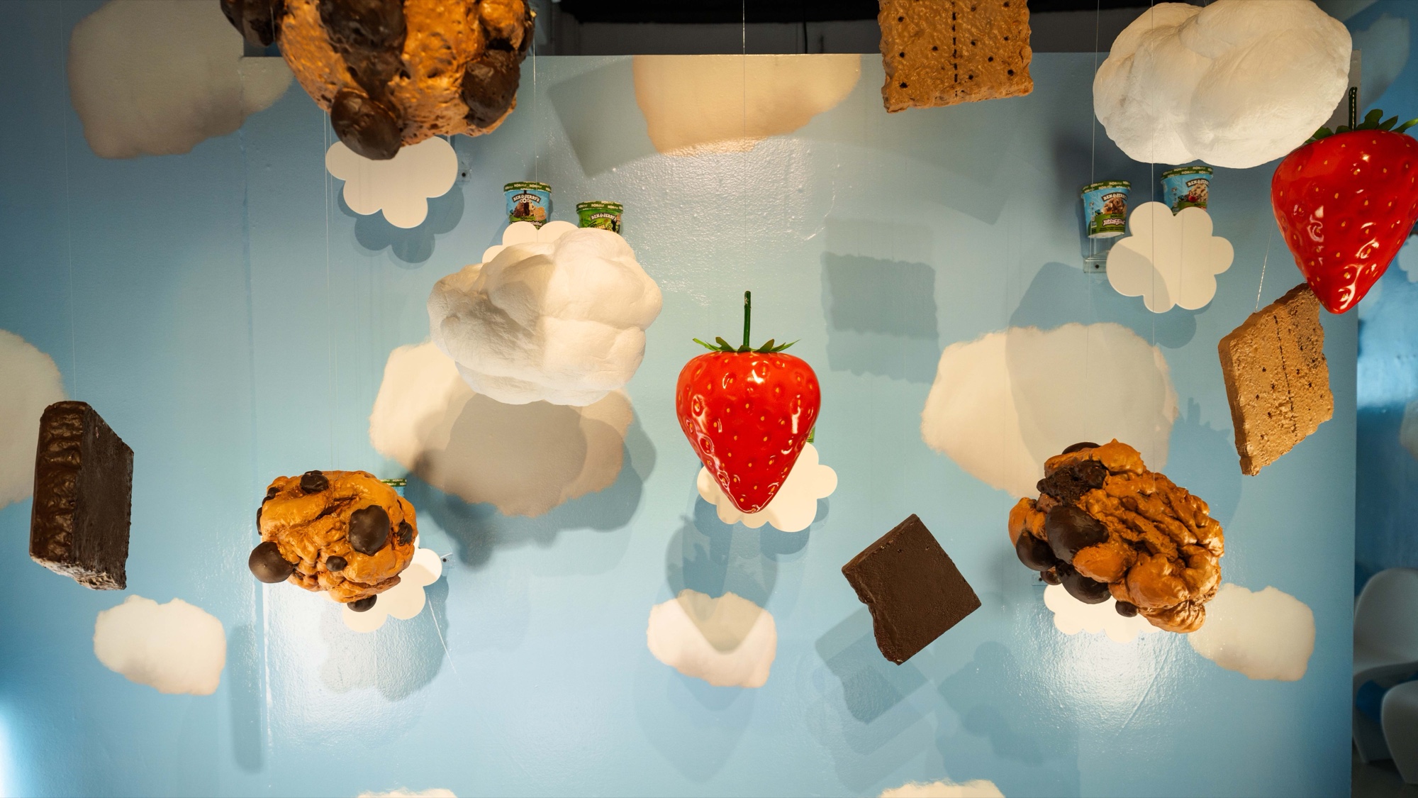 Giant cookies, brownies and strawberry hang with clouds and Ben & Jerry’s pints on a sky blue wall