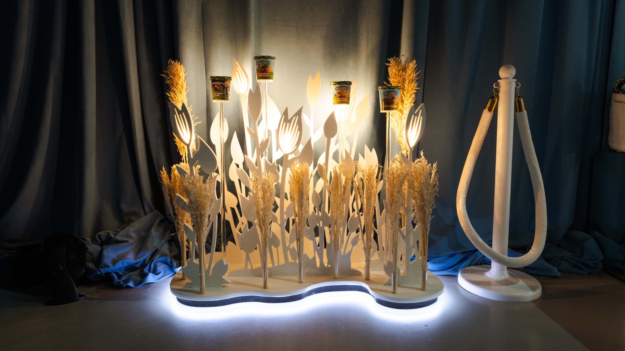 Warmly lit installation of Ben & Jerry’s pints atop white botanical cutouts and wheat with a glowing base