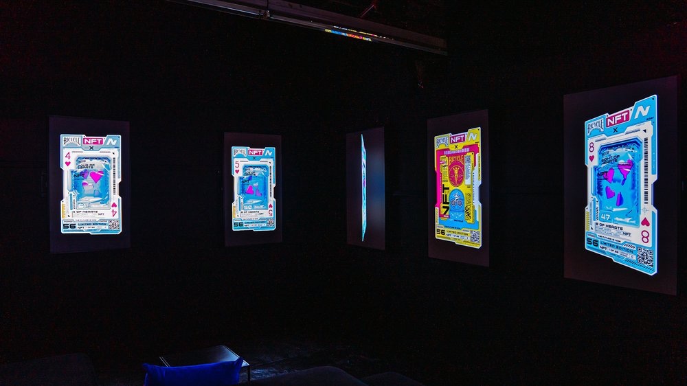 Glowing digital card screens from the Bicycle NFT collection line a darkened gallery
