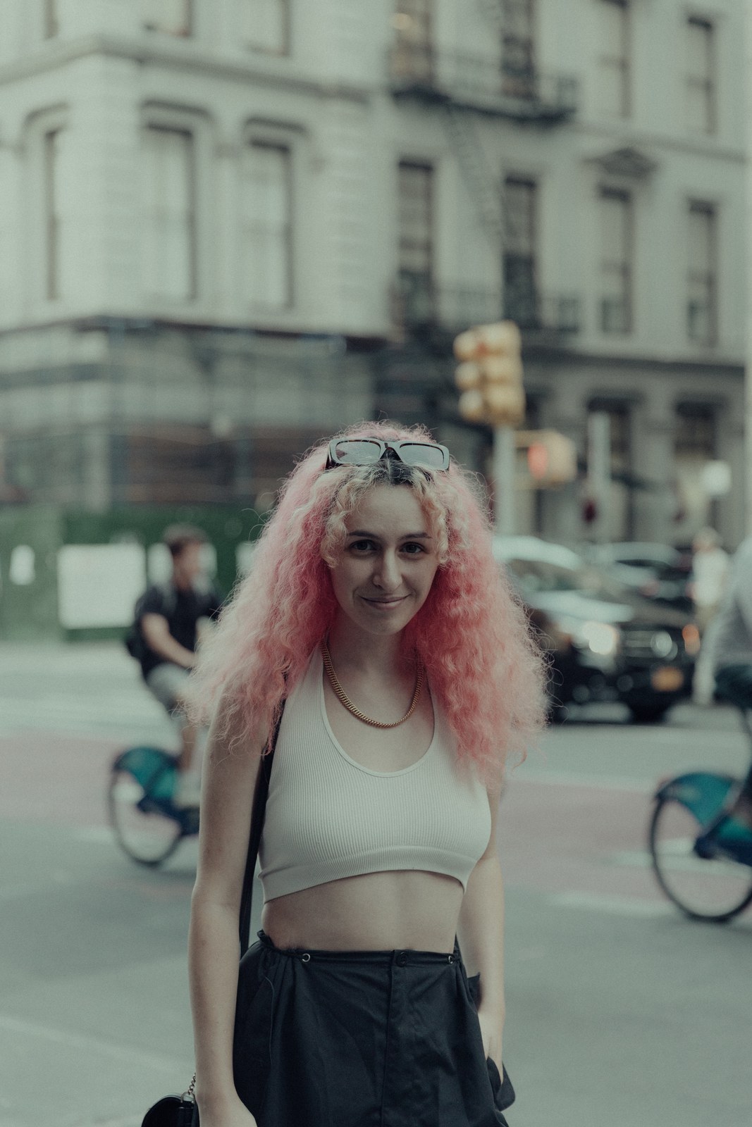 Woman with long pink curls and a white crop top smiles on a city crosswalk with blue share bikes behind