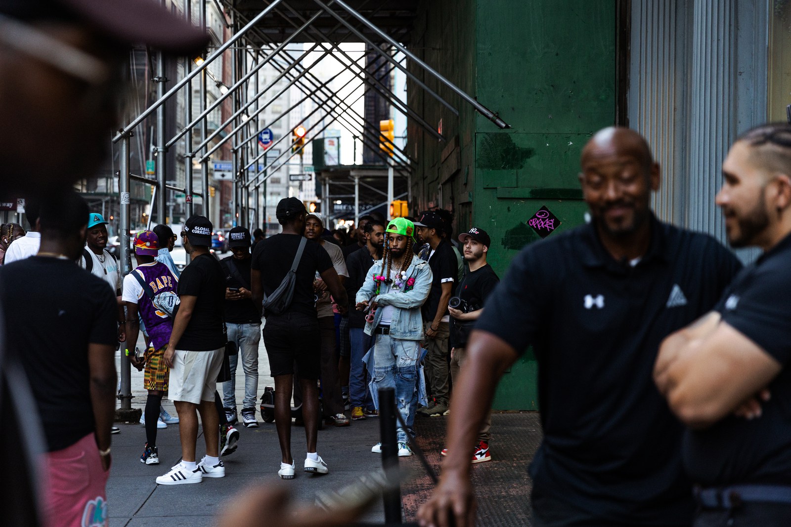 Crowd waits on a Manhattan sidewalk under scaffolding while two men in black polos stand in the foreground
