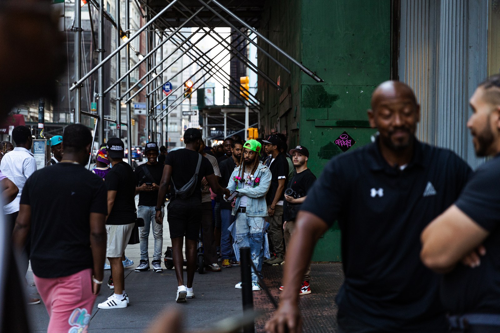 Crowd waits under sidewalk scaffolding beside a green building while two men in black polos stand in front