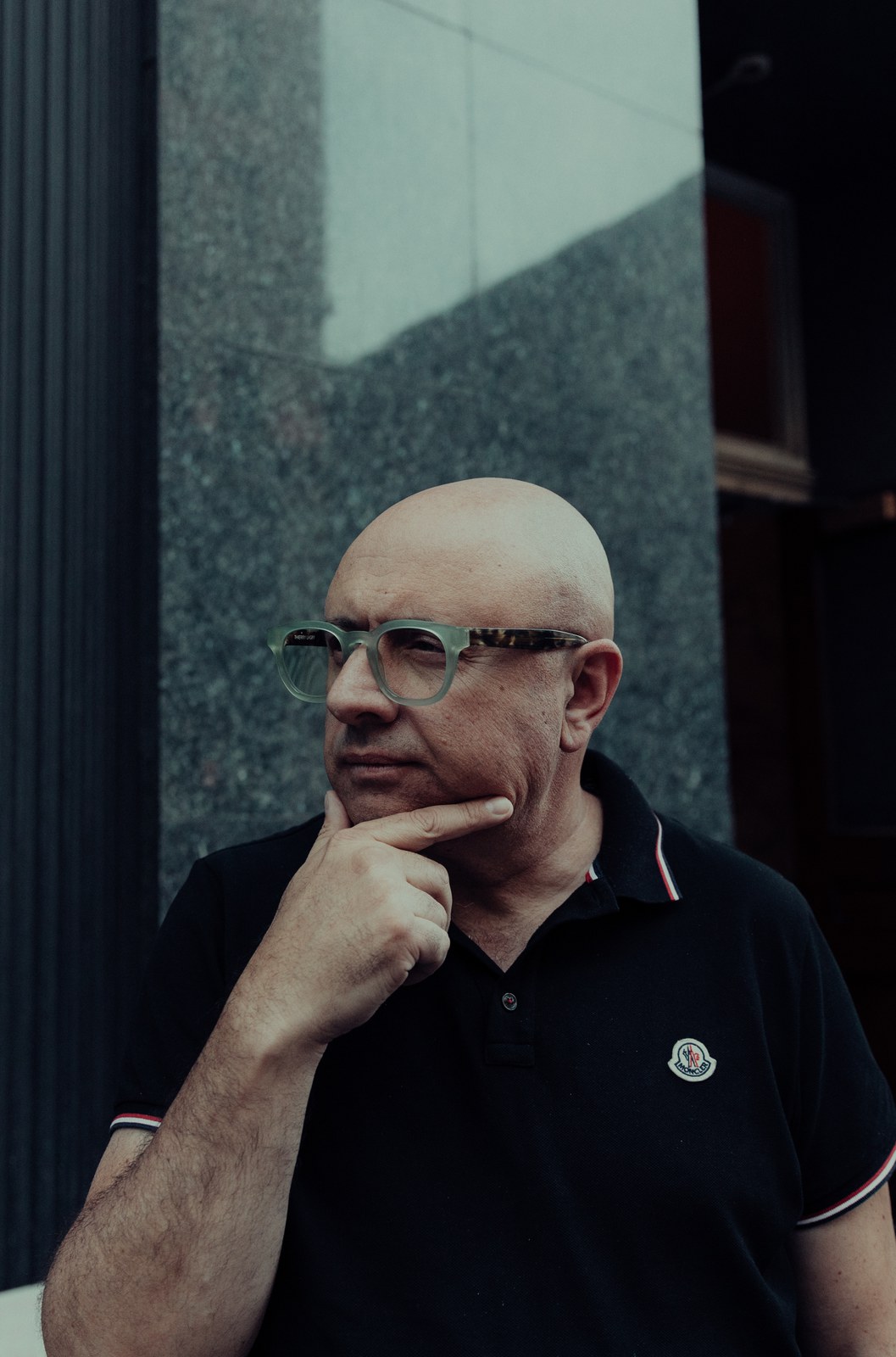 Bald man in green framed glasses and black polo rests his chin on his hand outside a granite facade