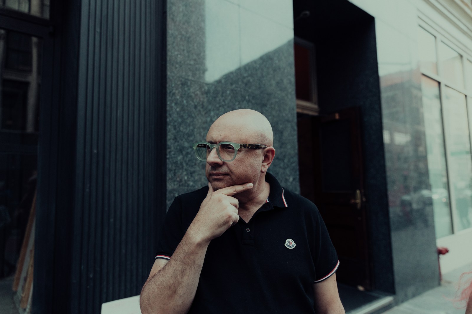 Bald man in clear framed glasses and a black polo rests his chin on his hand outside a stone building