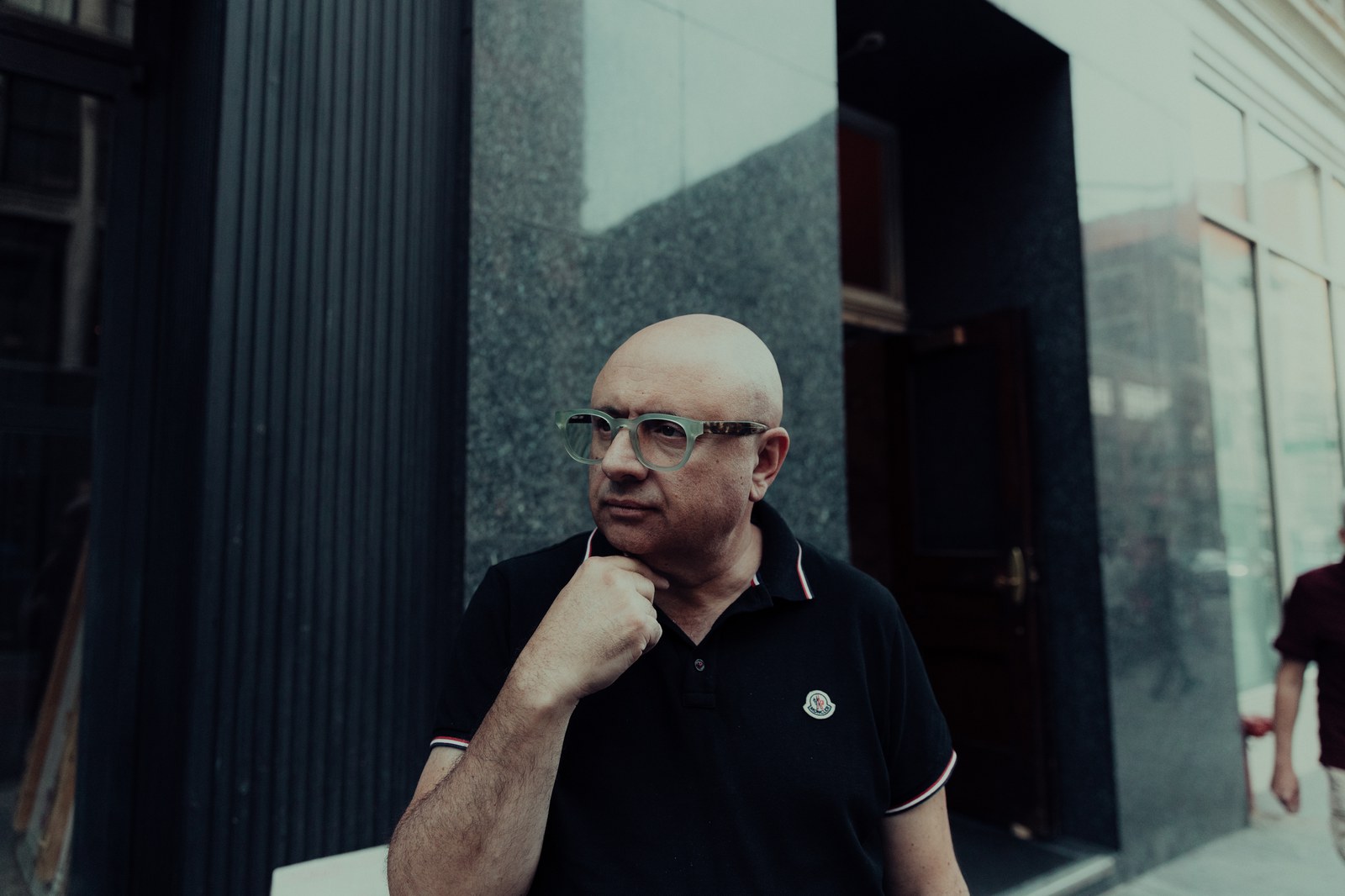 Bald man with clear framed glasses in a black polo, hand on chin, outside a dark stone building