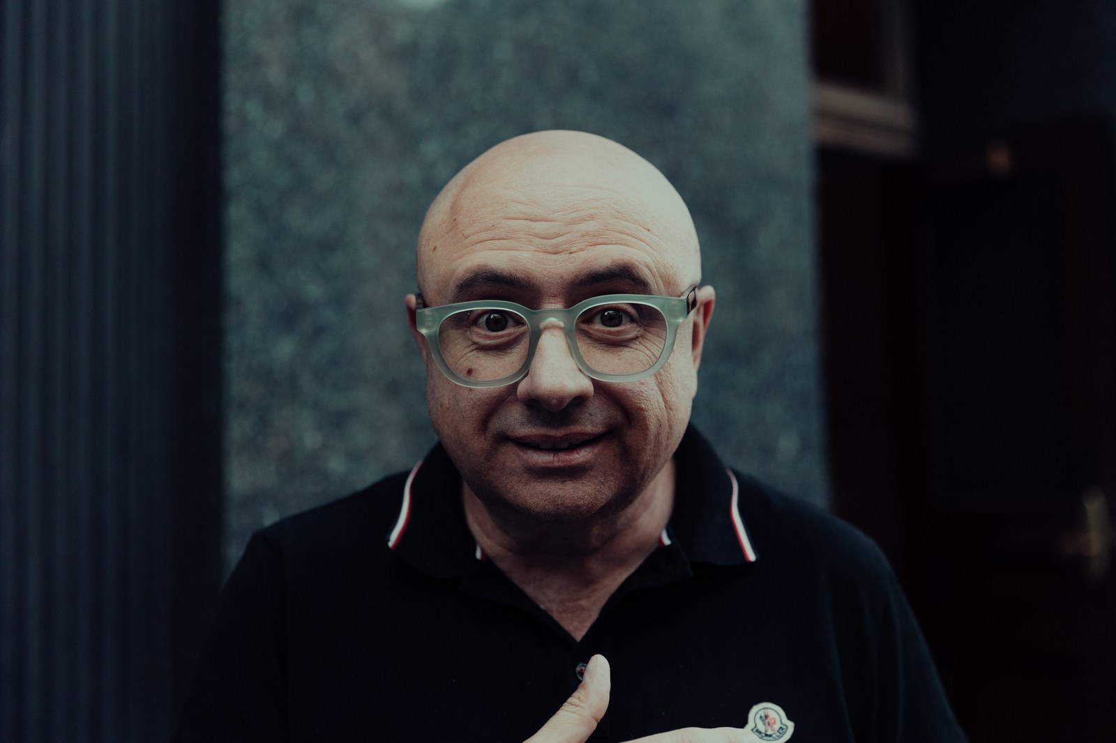 Bald man in green glasses smiles at the camera with a hand on his chest, dark facade behind