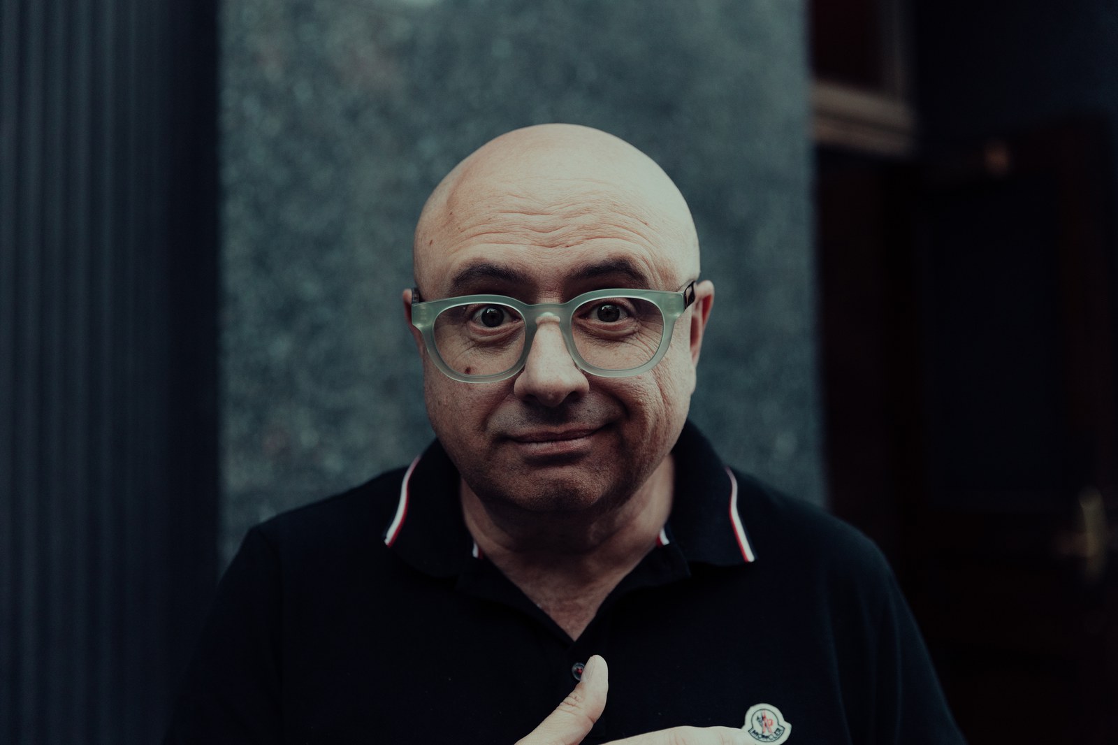 Bald man in clear framed glasses and a black polo looks into the camera with a hand on his chest