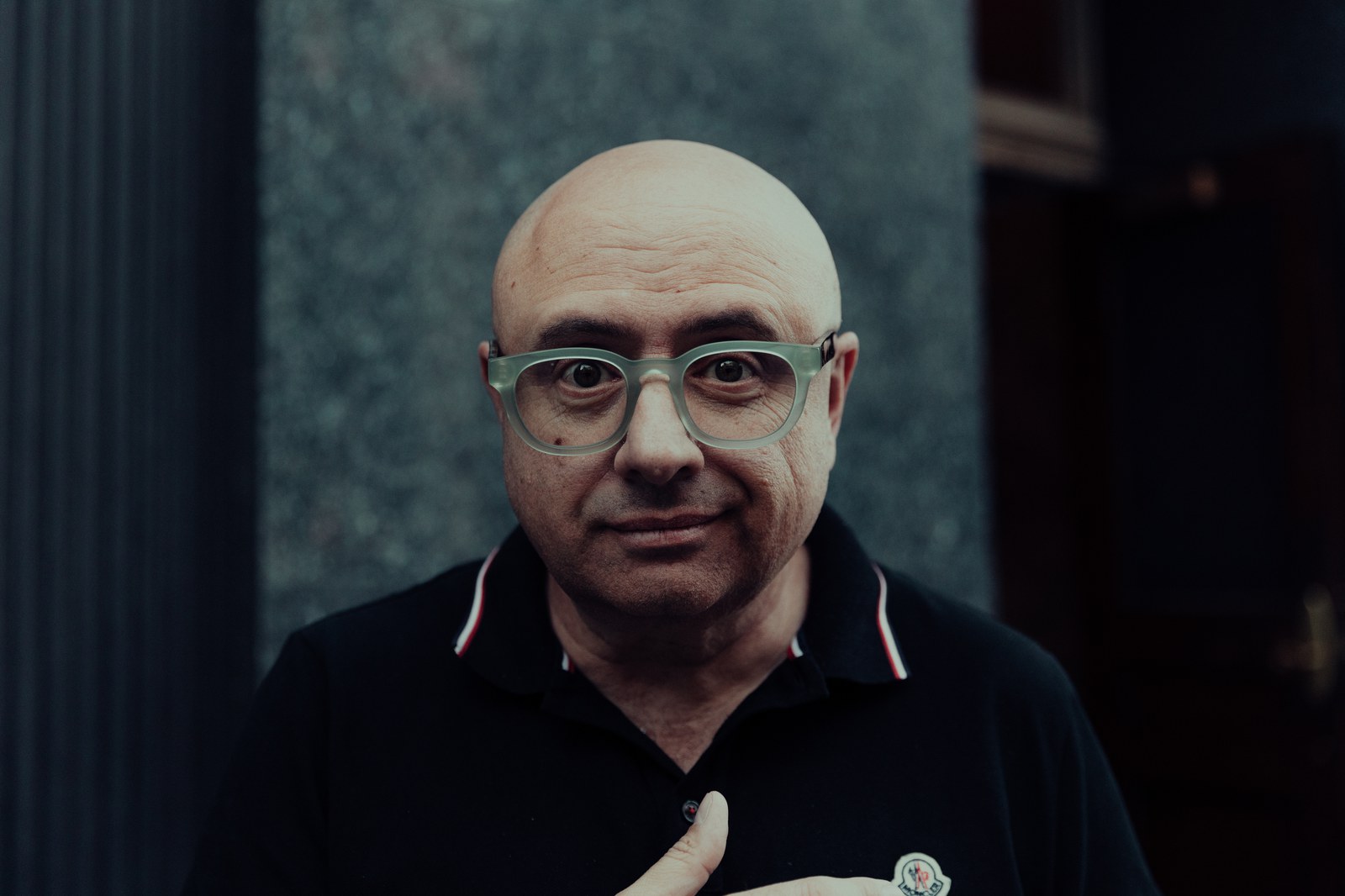 Close portrait of a bald man in clear glasses and a black polo with a hand on his chest