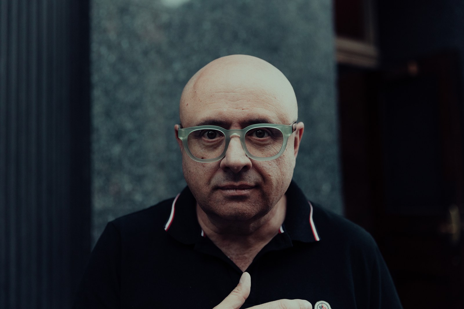 Close portrait of a bald man in green glasses looking straight at the camera with a hand on his chest