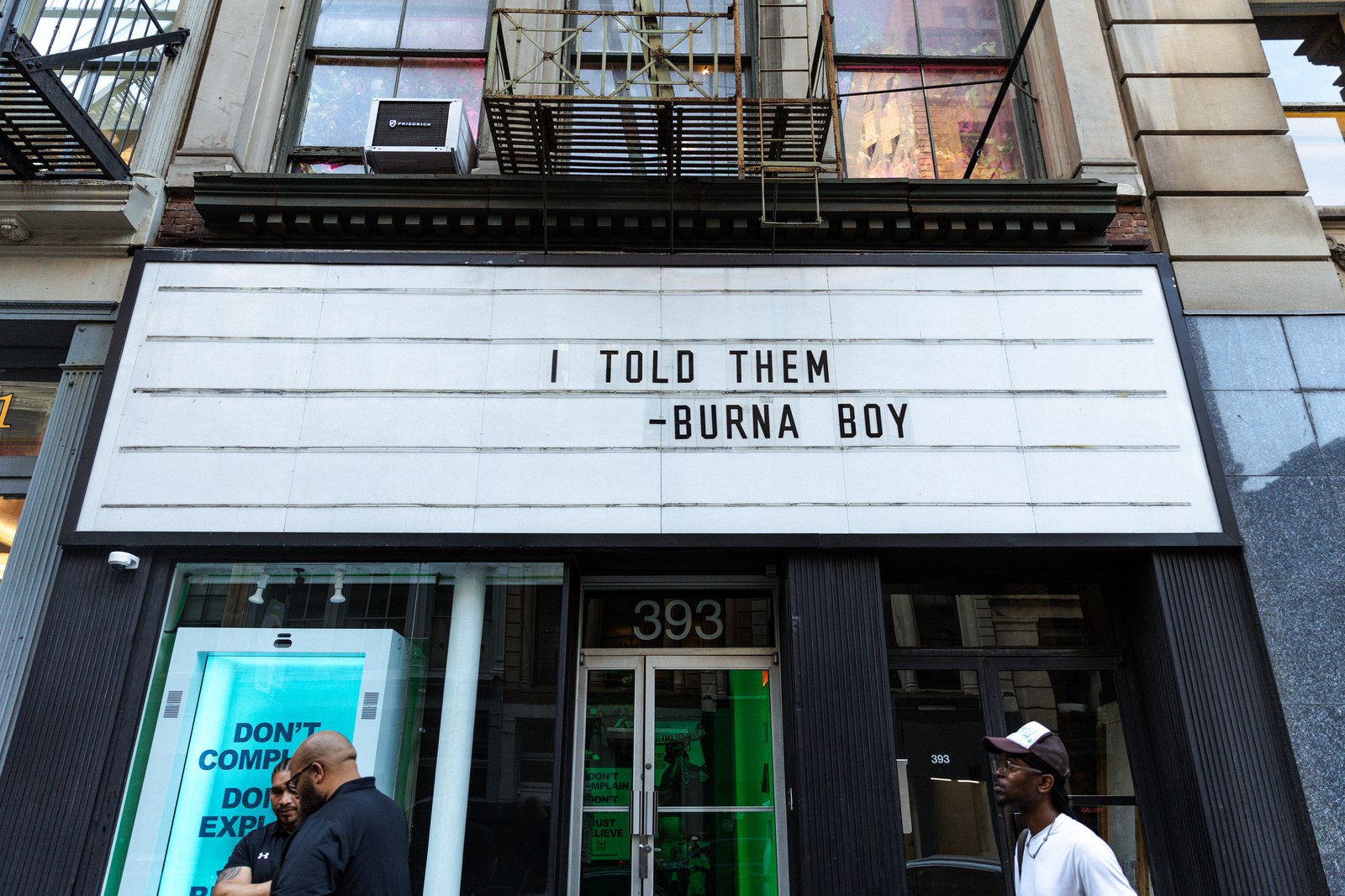 Marquee sign reading I Told Them Burna Boy above a storefront with fire escapes and a green window display