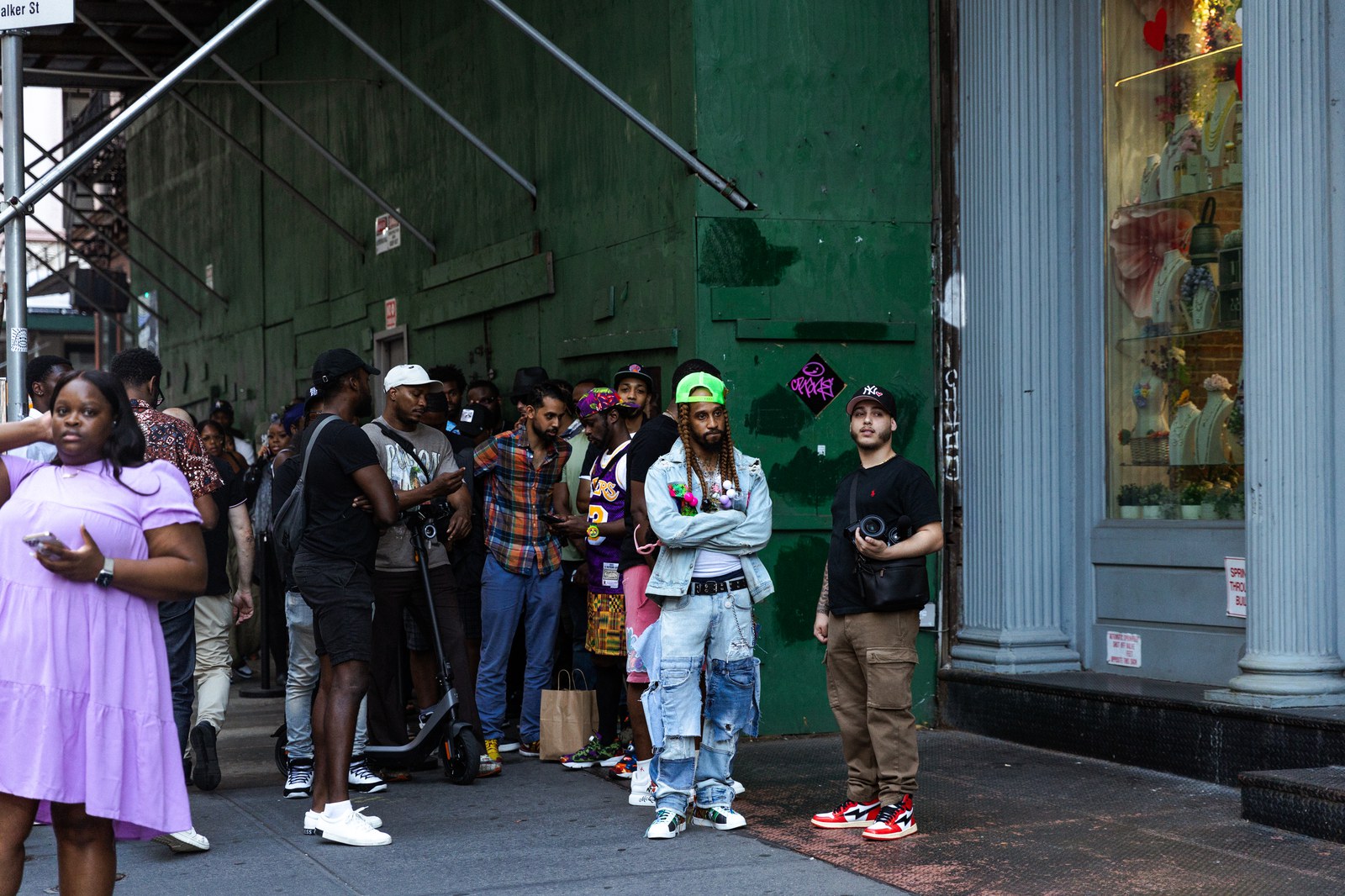 Line of people wait along a green wall on a city sidewalk, a woman in lilac at the front