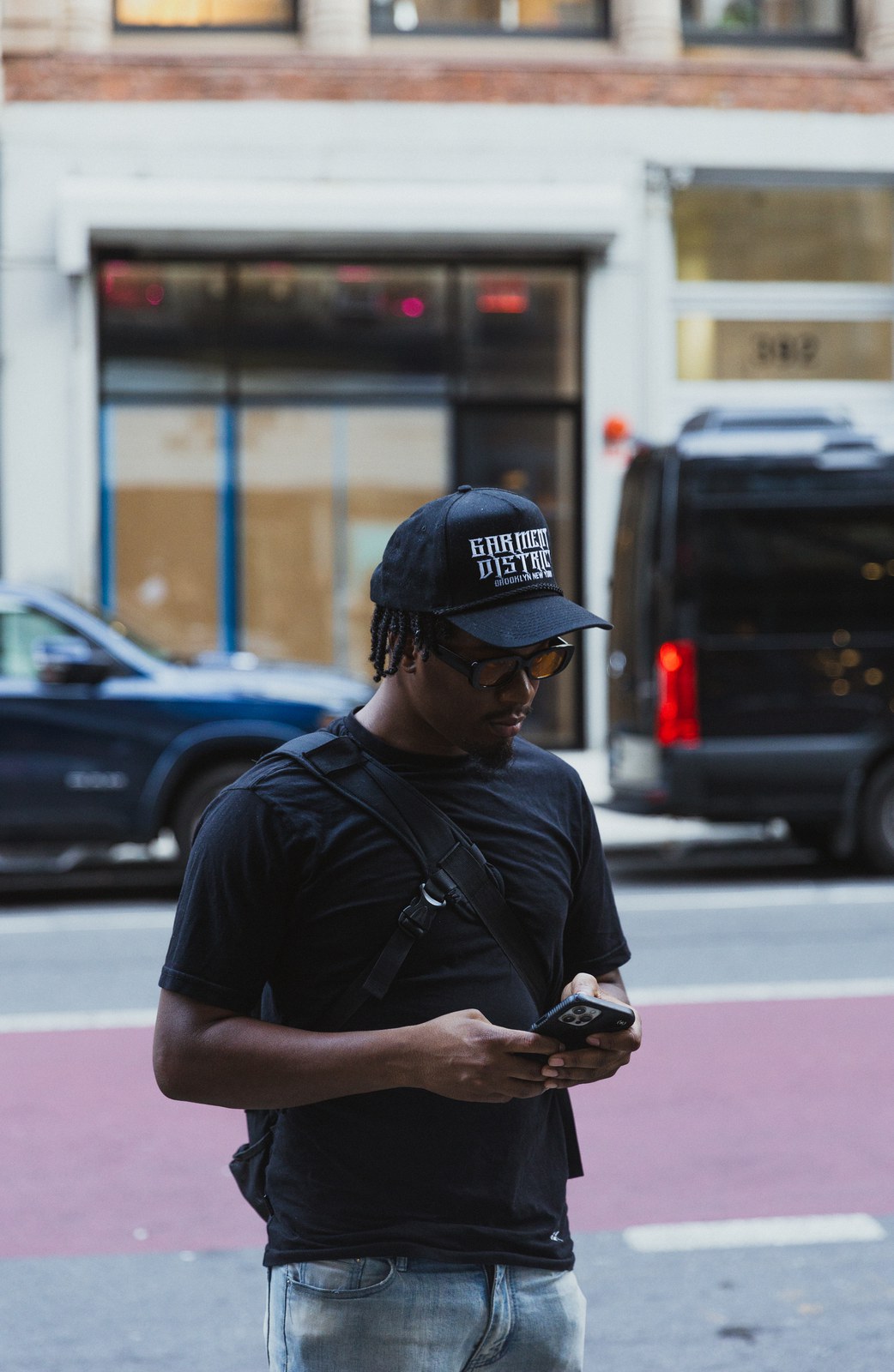 Man in a black cap and sunglasses checks his phone on a street corner with a van parked behind