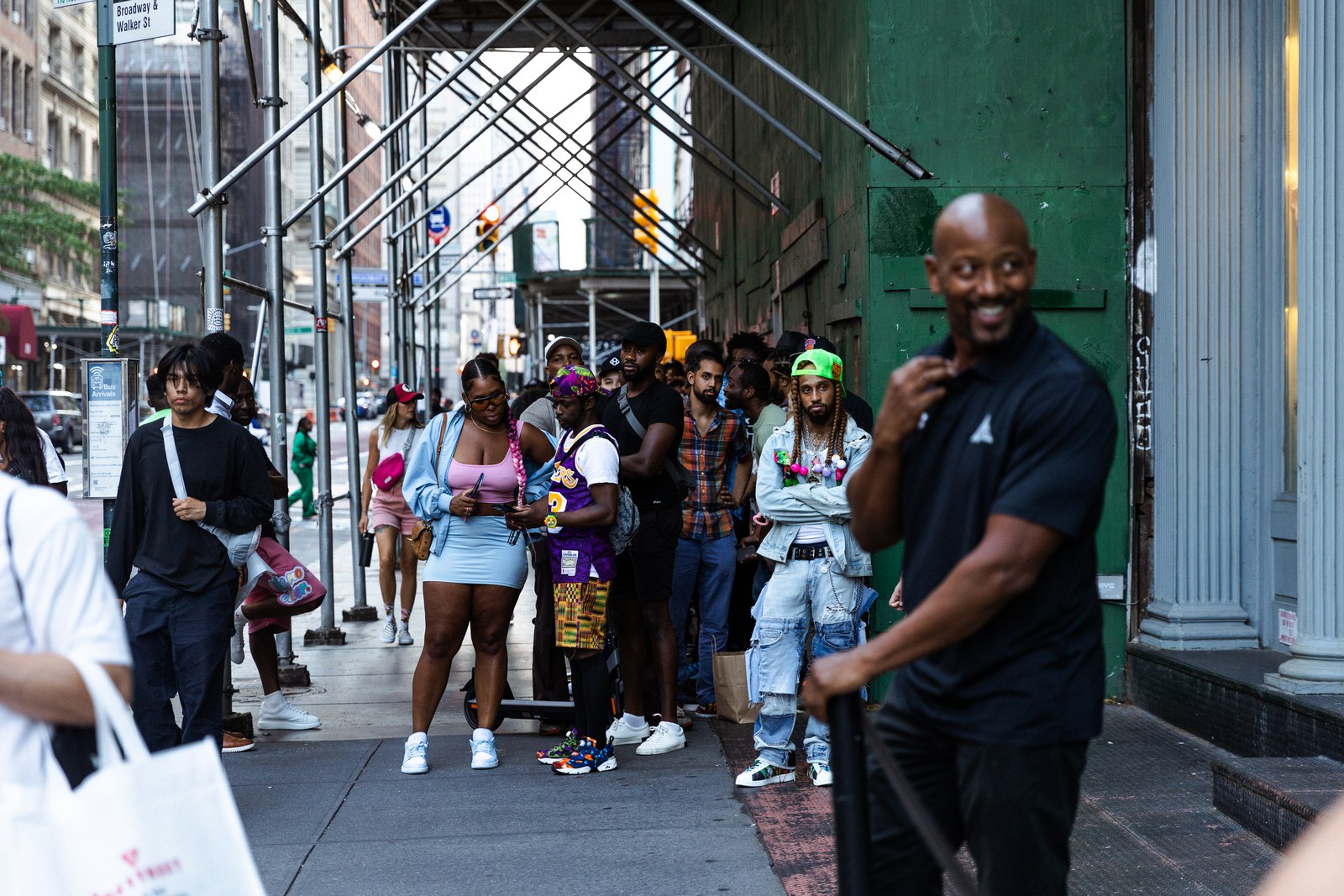 Line of people waits under sidewalk scaffolding as a smiling man in black walks past in the foreground