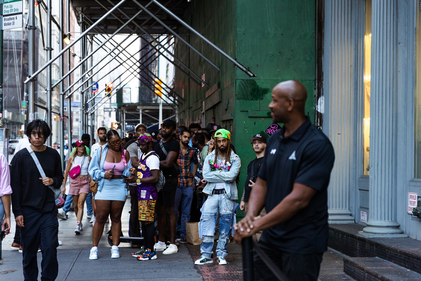 Queue under scaffolding beside a green building as a man in a black polo stands at the corner