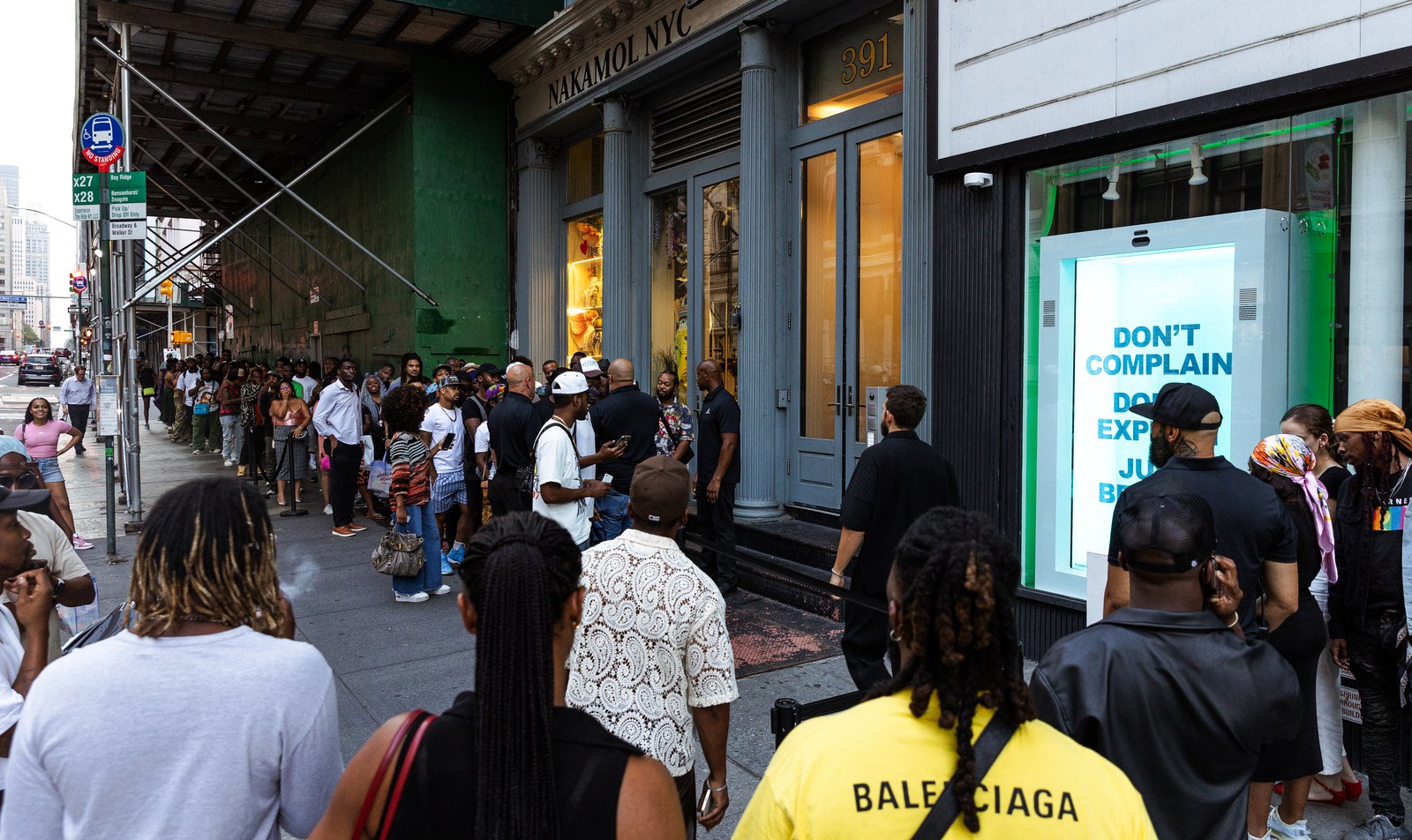 Crowd lines a city sidewalk outside a storefront with a Don’t Complain sign in the window
