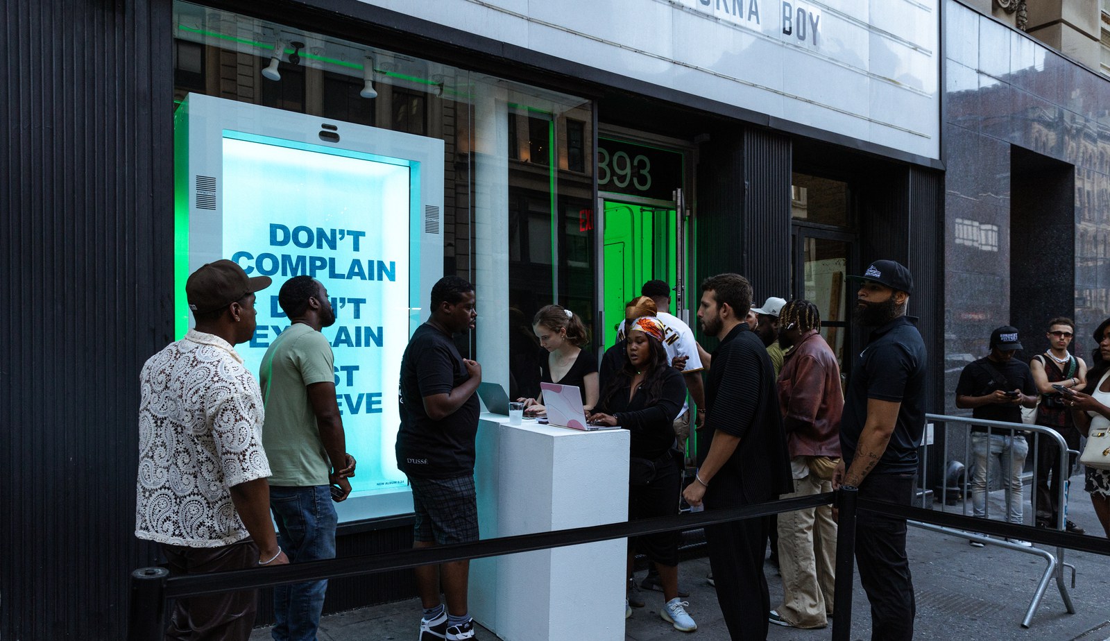 Guests queue at a white check in desk outside a storefront window screen reading Don’t Complain