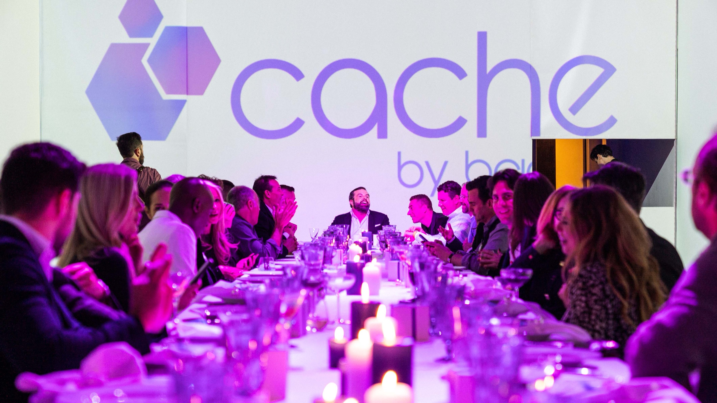 Guests applaud along a candlelit dinner table beneath a projected Cache logo in purple light
