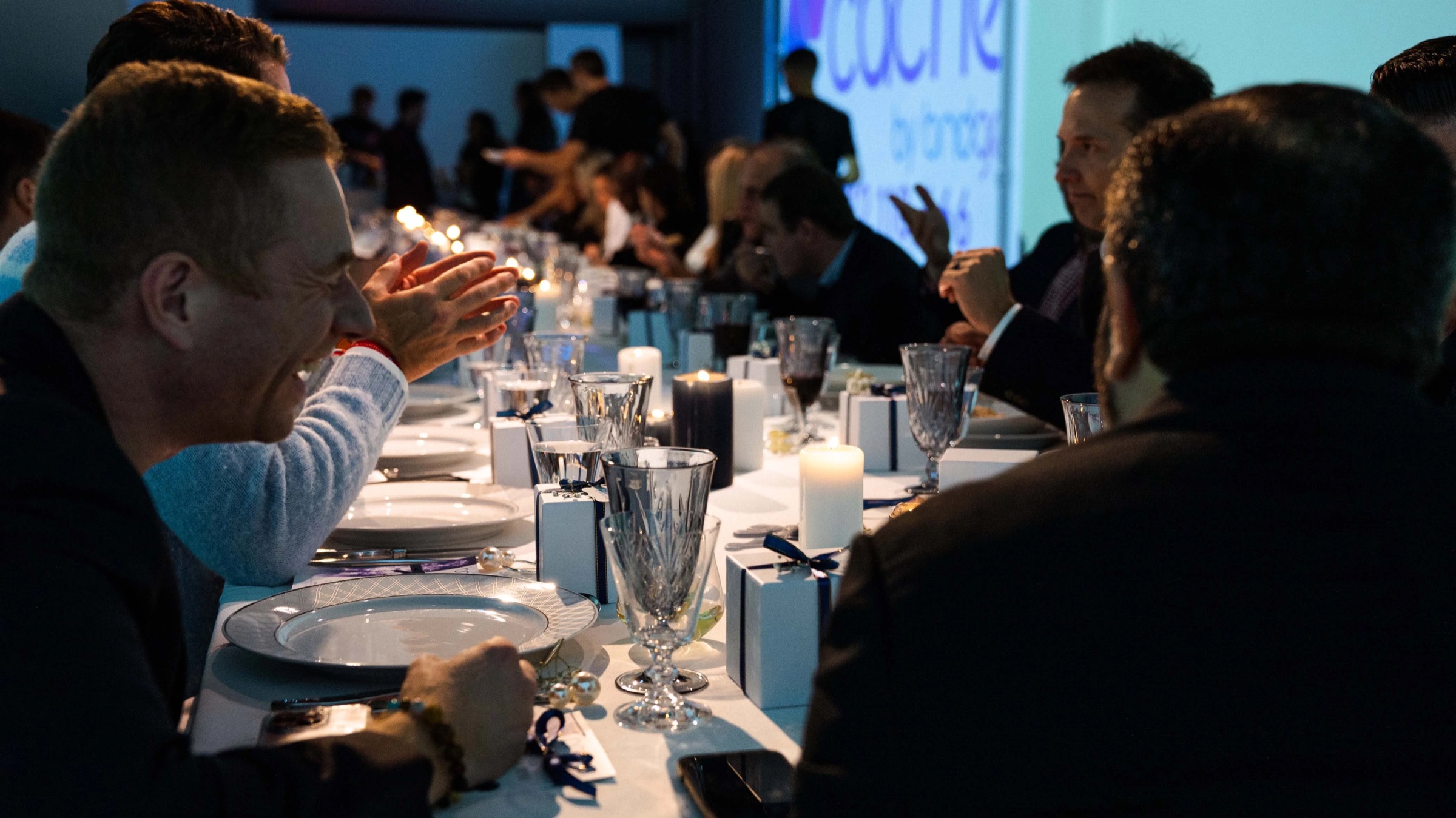 A guest laughs across a candlelit dinner table with the Cache by Bridge logo projected behind him