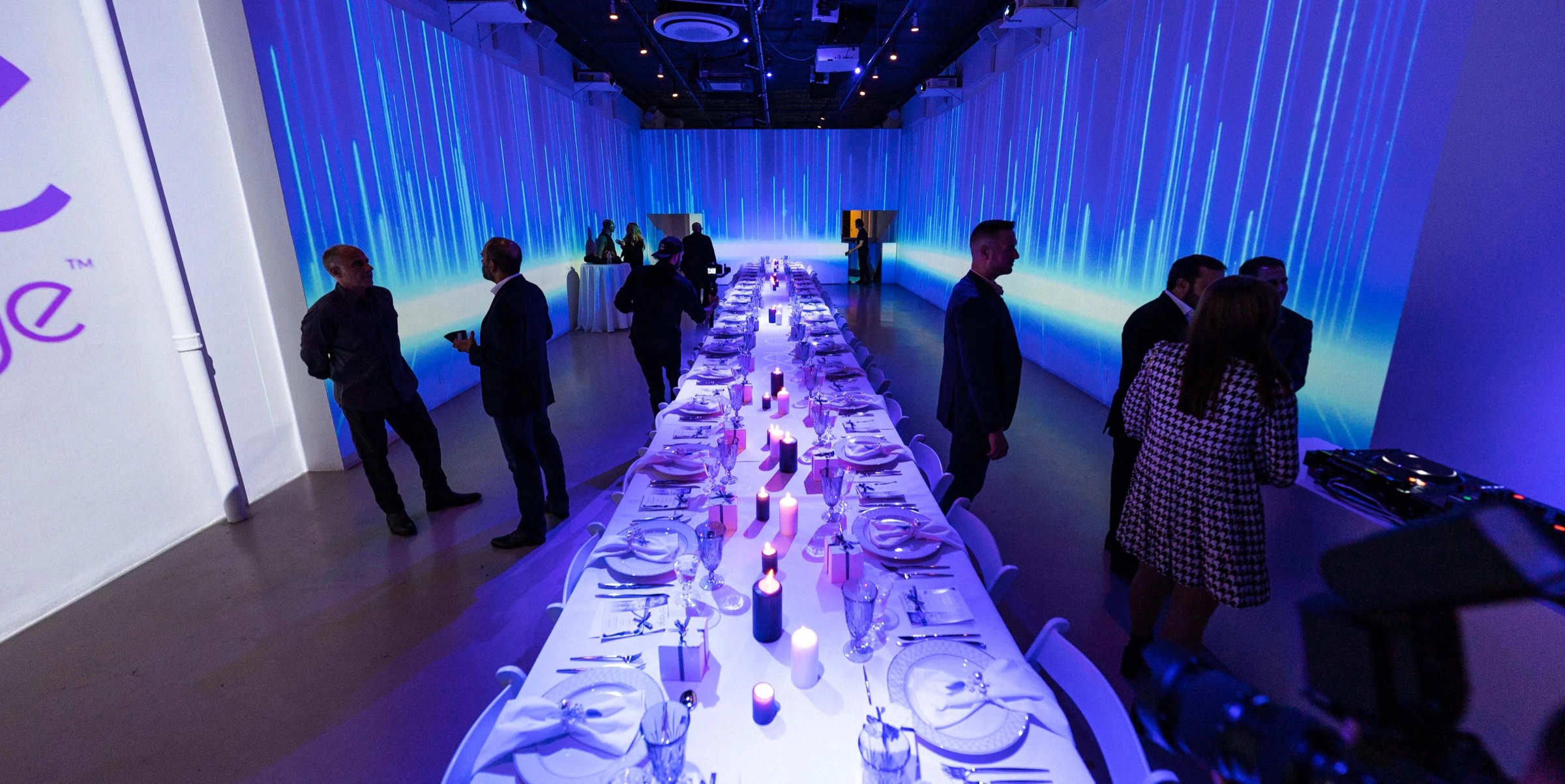 Overhead view of a long white dinner table lit in violet beneath streaked blue light projections at the Cache by Bridge dinner
