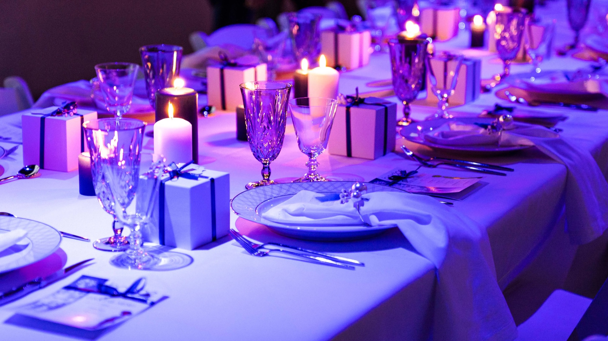Place setting with a white gift box, pillar candles and crystal glassware washed in violet light