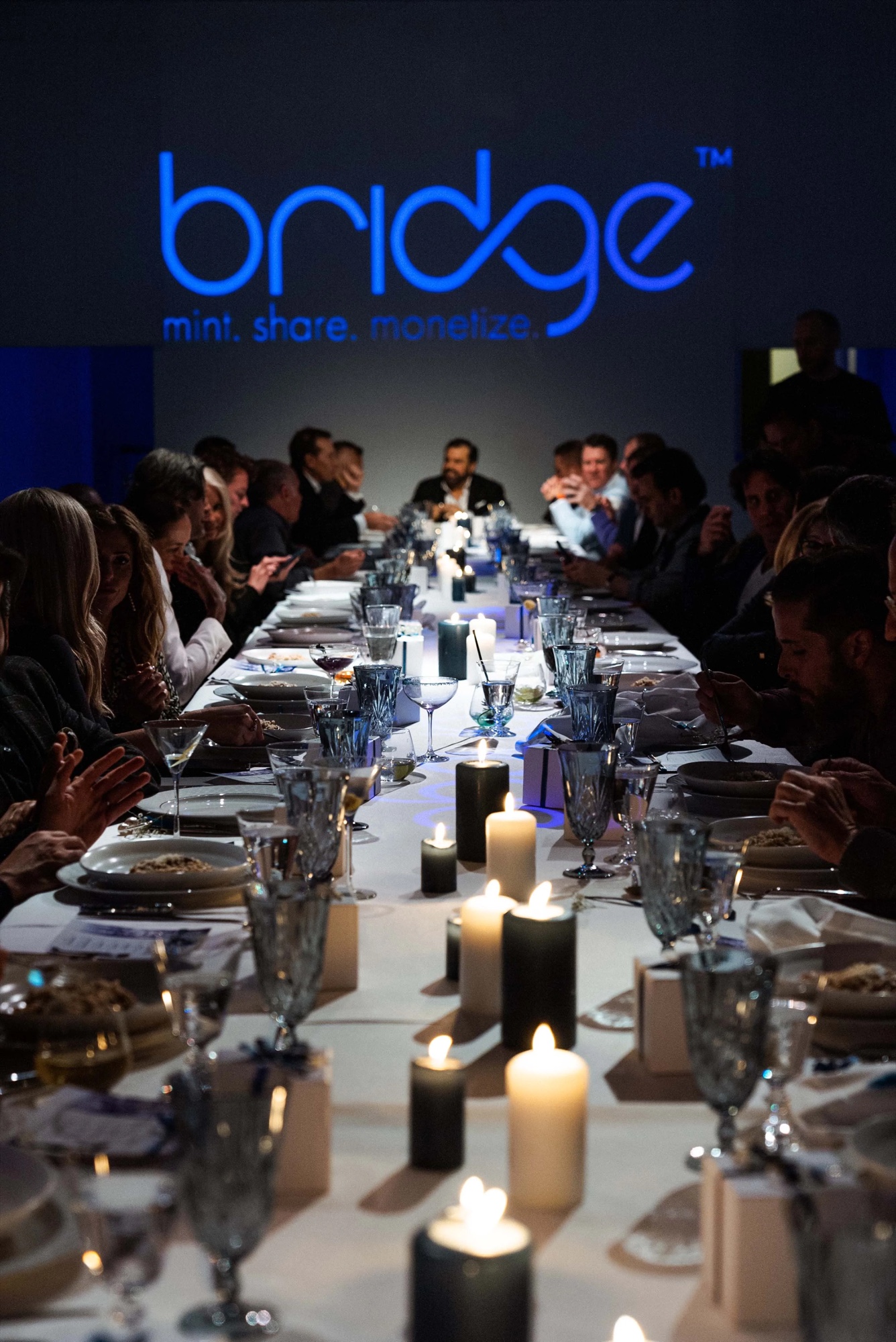 Diners seated beneath a projected Bridge logo reading mint, share, monetize at Lume Studios