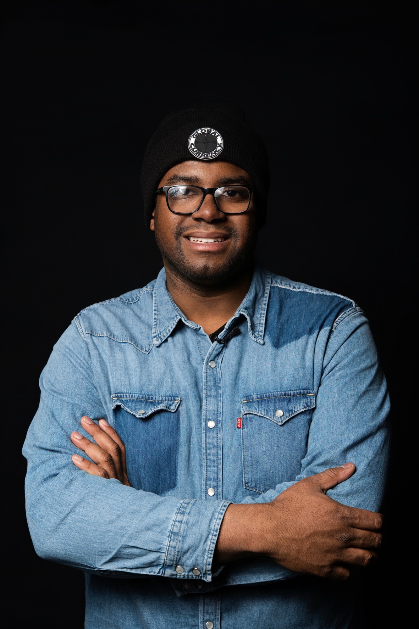 Kyle Bethel in a denim shirt and black beanie, arms folded, smiling toward the camera
