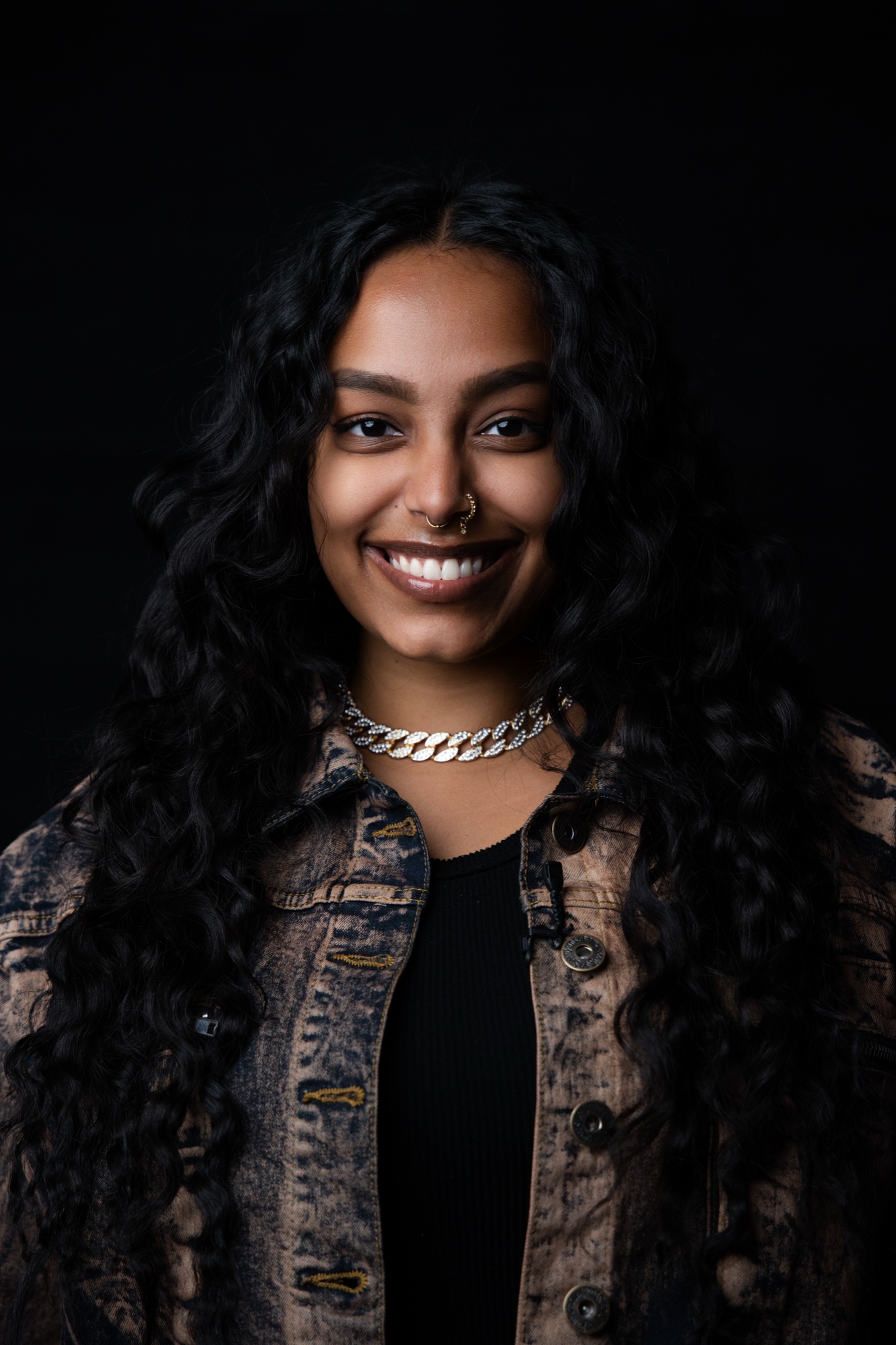 Subiy smiling in a washed denim jacket over a black top, silver chain at the neck