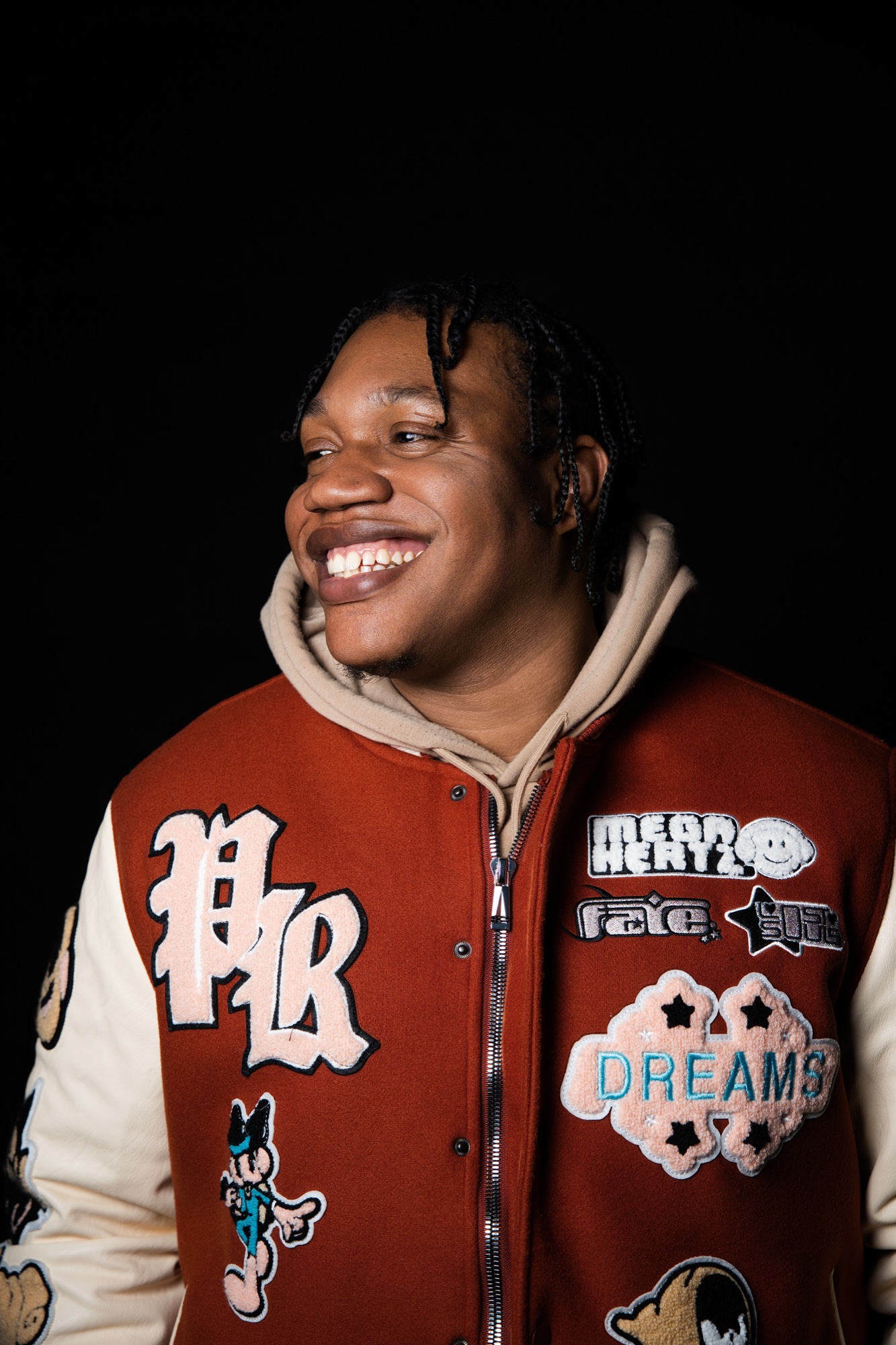 Drama B laughing in a red varsity jacket patched with DREAMS, looking off to one side