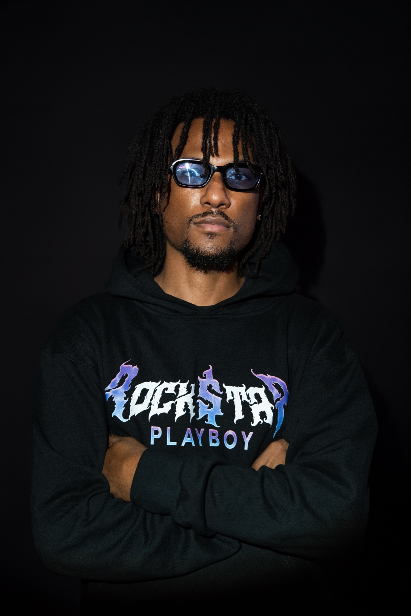 FritzThaProducer with arms folded in a black ROCKSTAR PLAYBOY hoodie and dark sunglasses