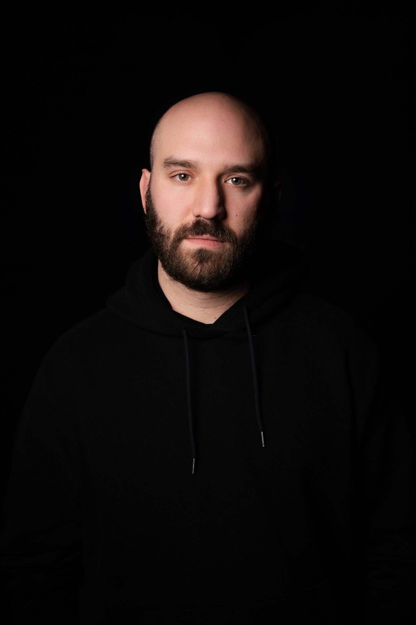 Ari Axelrod in a plain black hoodie, bearded, looking straight at the camera under low light