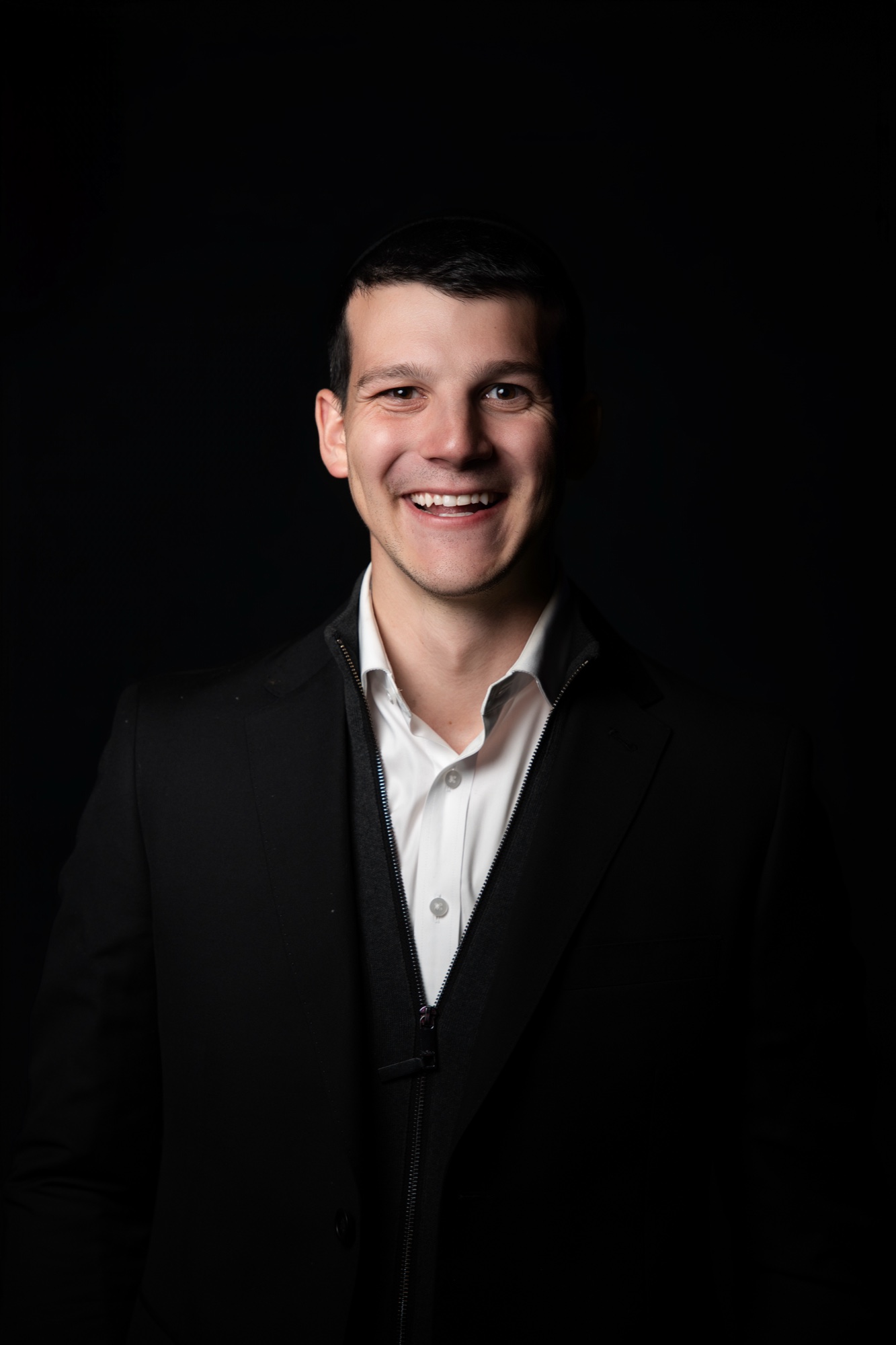 Tai smiling in a black jacket over a white collared shirt, lit from the left