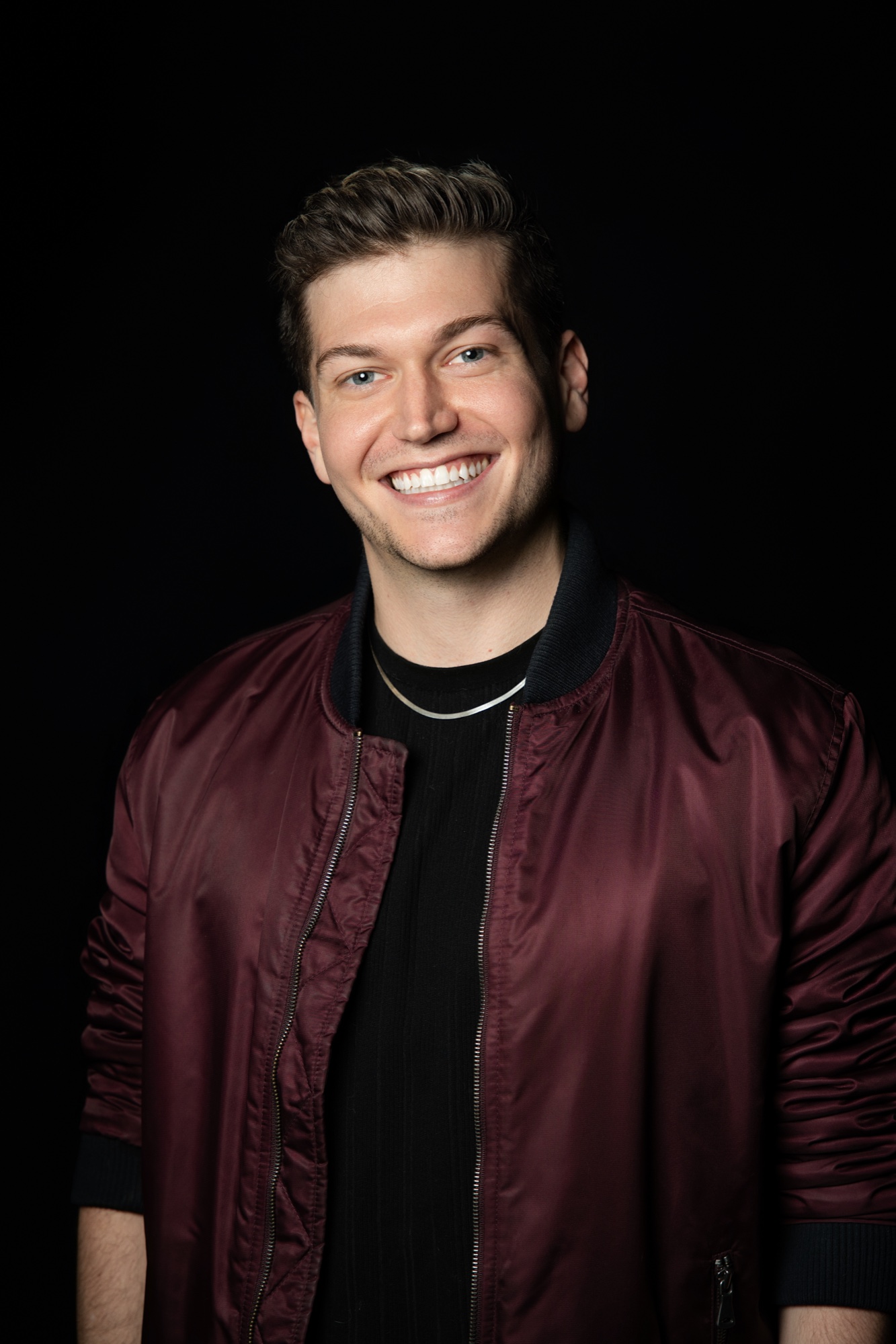 Tyler Palo grinning in a maroon bomber jacket over a black tee with a thin chain