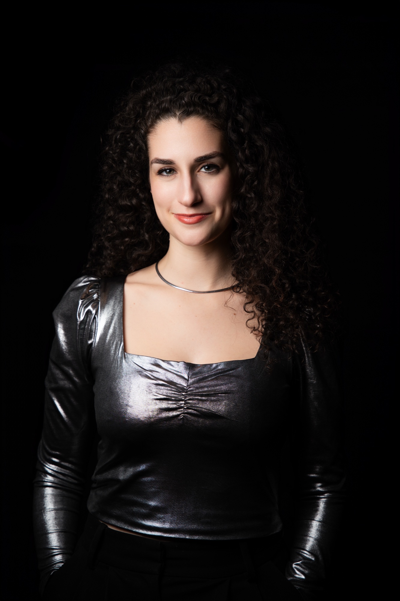 Izzy Stein in a silver metallic top and thin necklace, hands behind her back, smiling slightly