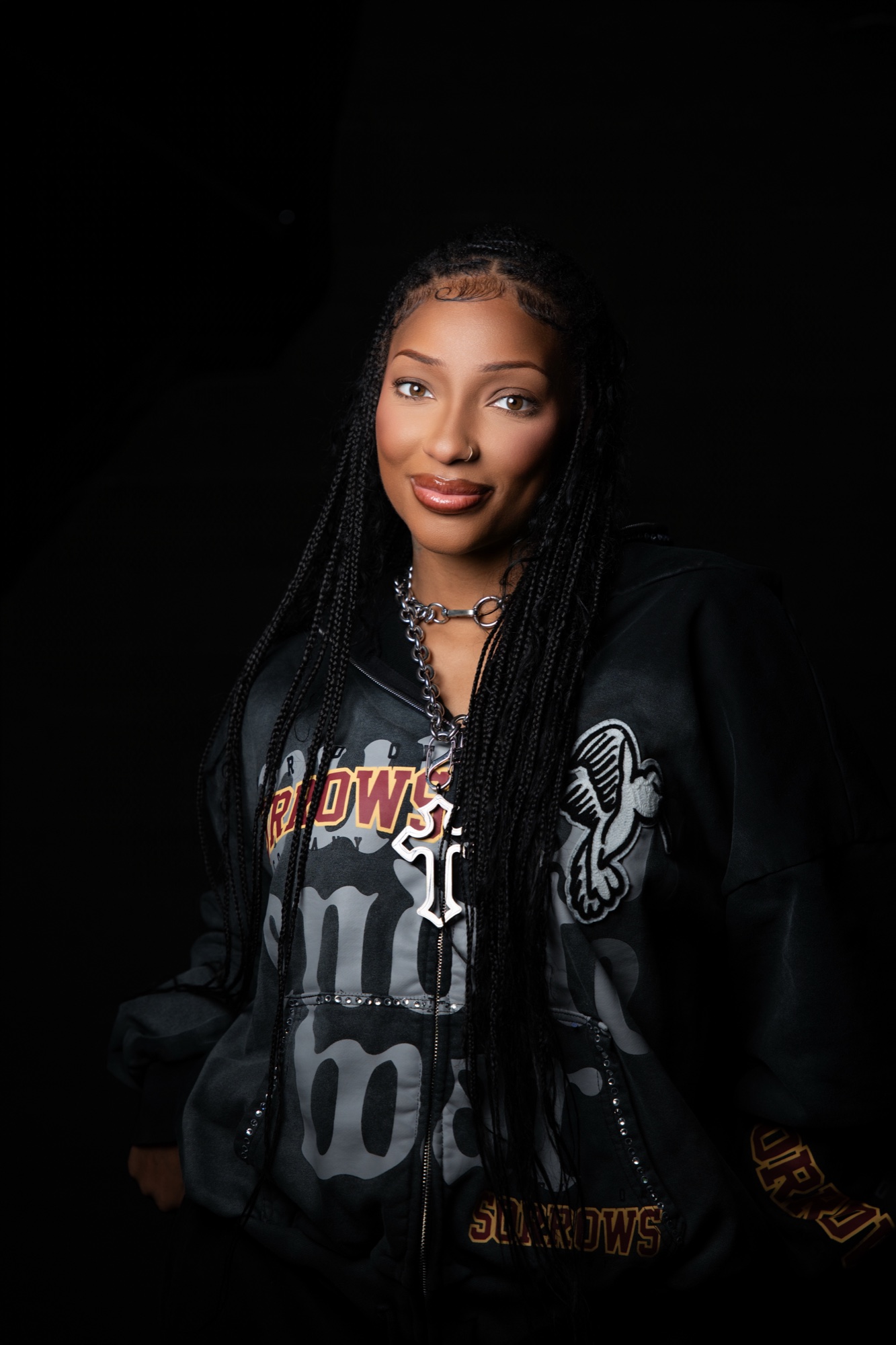 LeA Robinson in a black graphic hoodie and silver chain, long braids, head tilted toward the camera