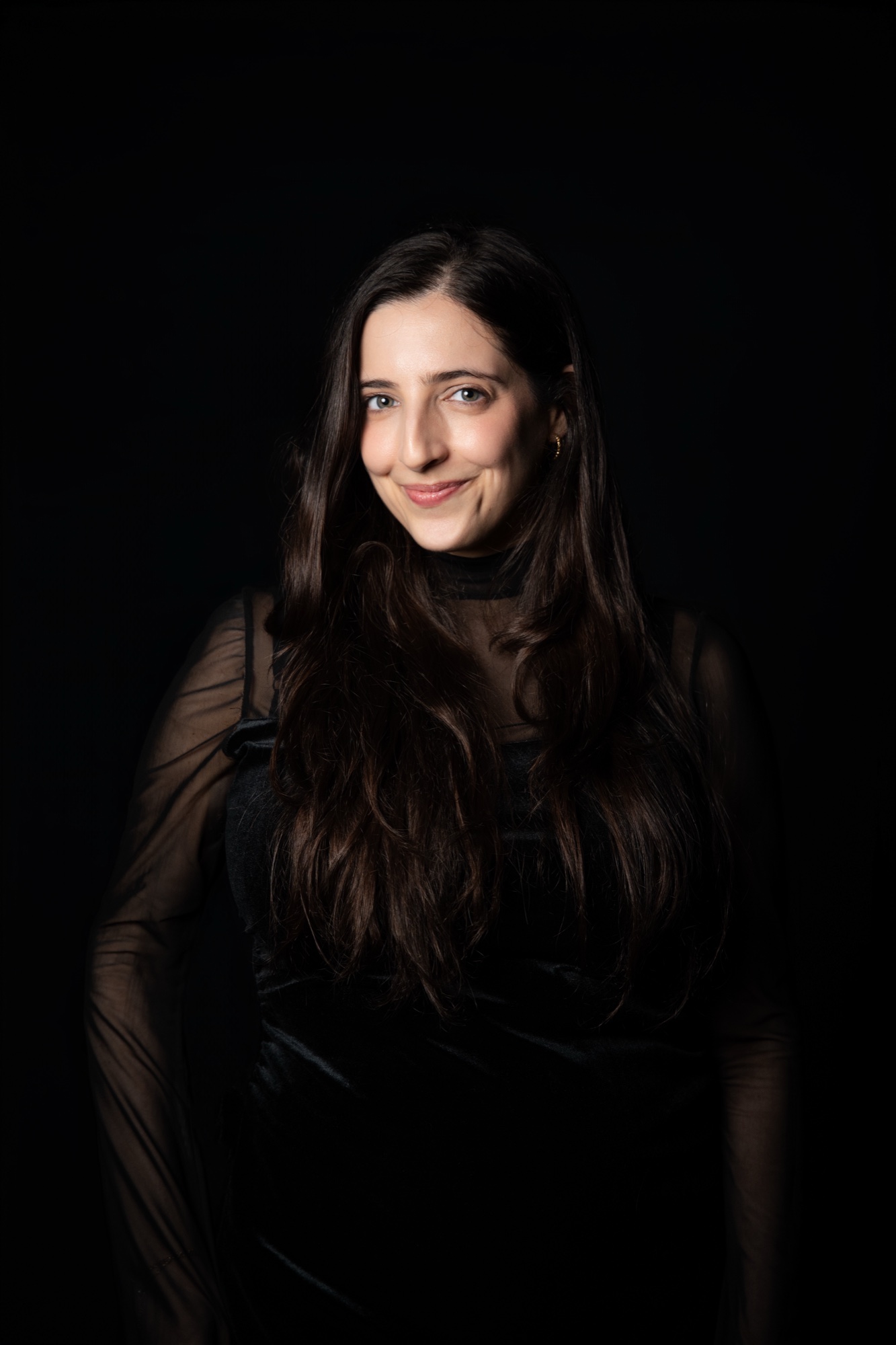 Tamar Sagiv in a sheer black top, long hair over one shoulder, smiling in dim light